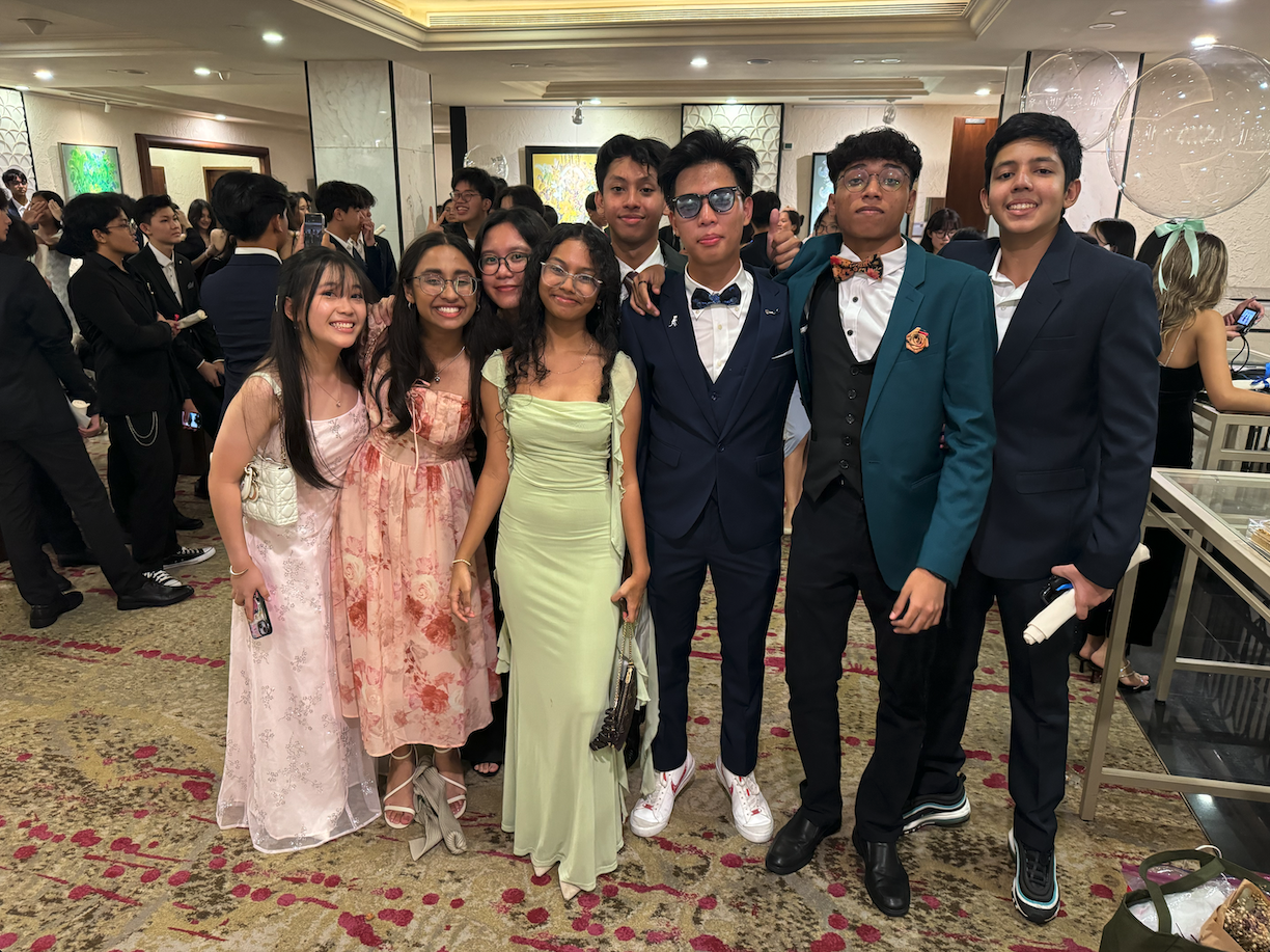 A group of seven teens in formal wear, including gowns and suits, posing together at a prom inside a ballroom.