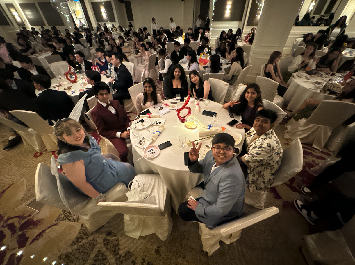 A large formal banquet hall filled with young people in formal attire seated at round tables with white tablecloths.
