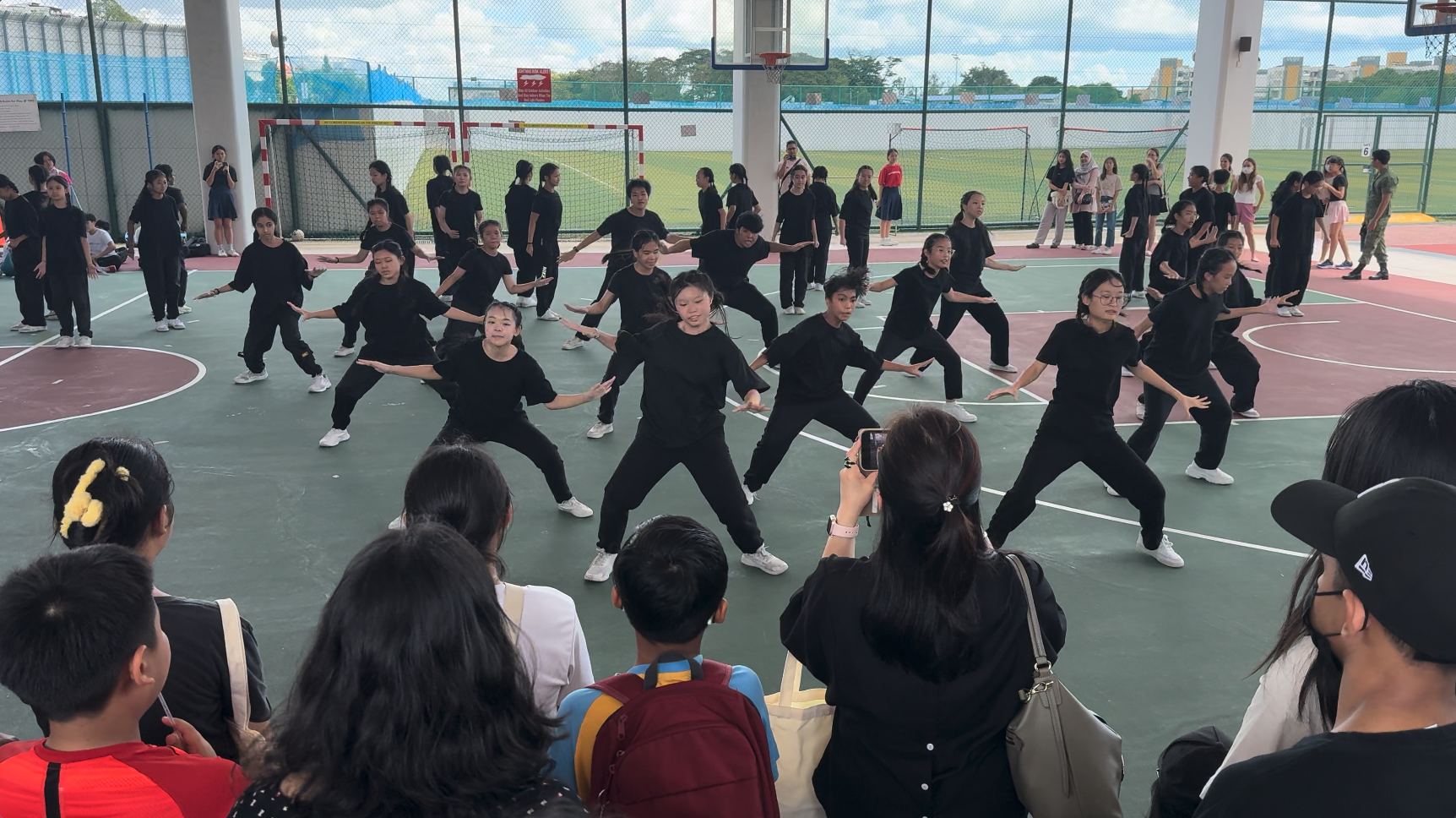 A large group of students in black outfits perform a synchronized dance on an outdoor basketball court before a crowd.