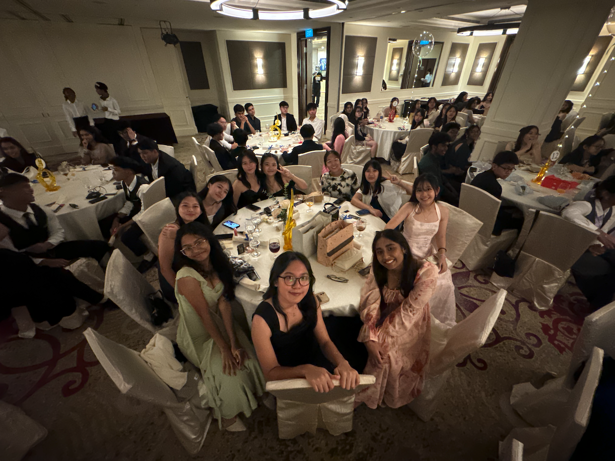 Young people in formal attire sit at round tables in a dimly lit banquet hall, smiling at the camera.