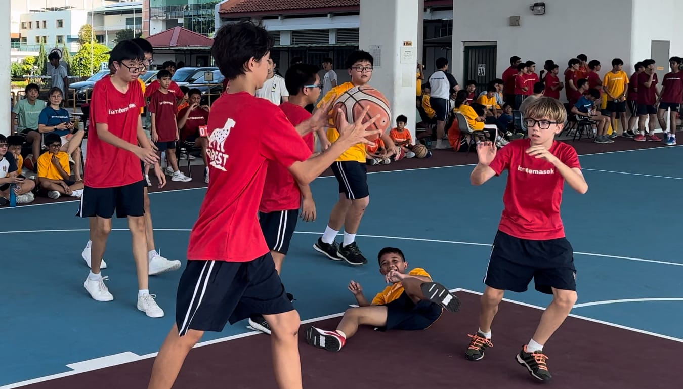 Boys in red and yellow jerseys play basketball on an outdoor court while spectators watch from the sides.