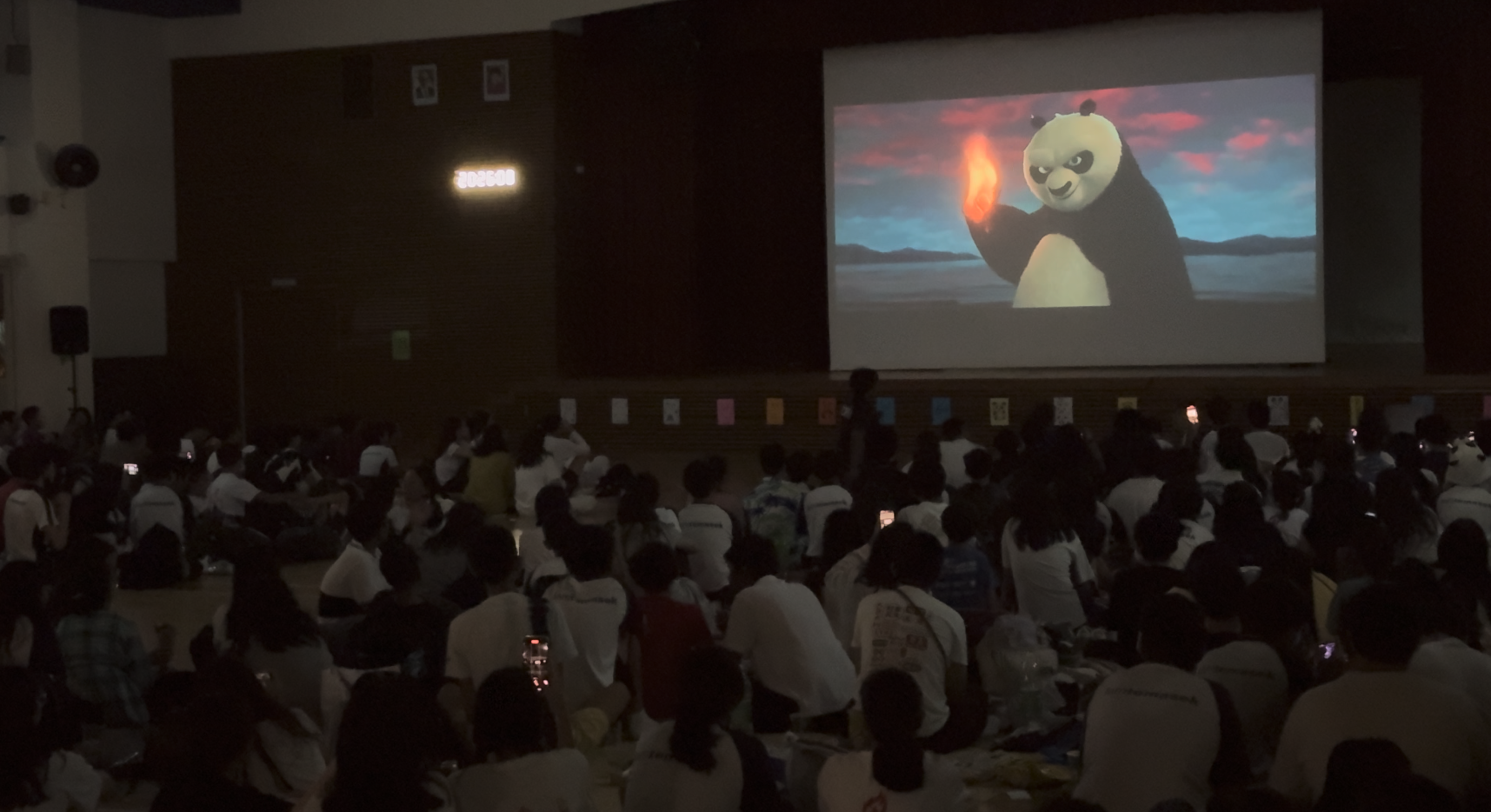 Large crowd seated in a dark hall watching Kung Fu Panda on a big projection screen.