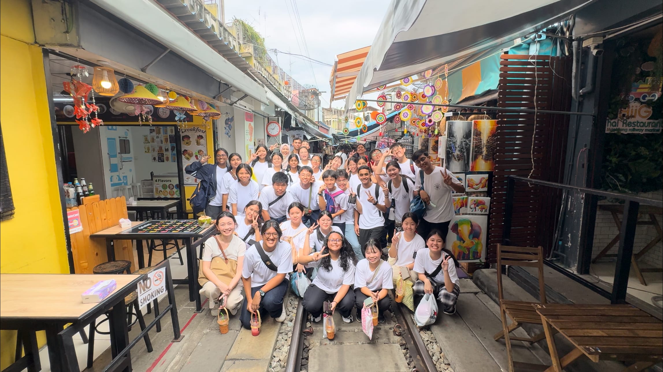 A large group of young people in white shirts pose smiling on a narrow market alley with railway tracks running through it.