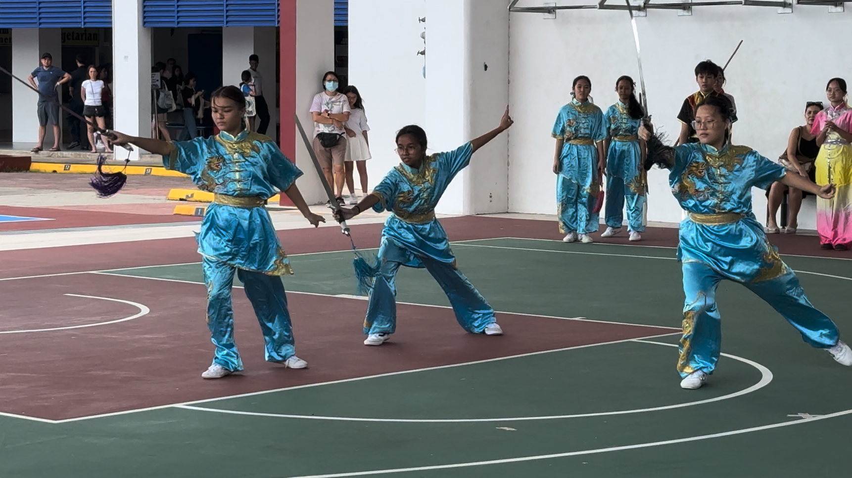 Young performers in blue and gold wushu uniforms wield swords on an outdoor court before spectators.