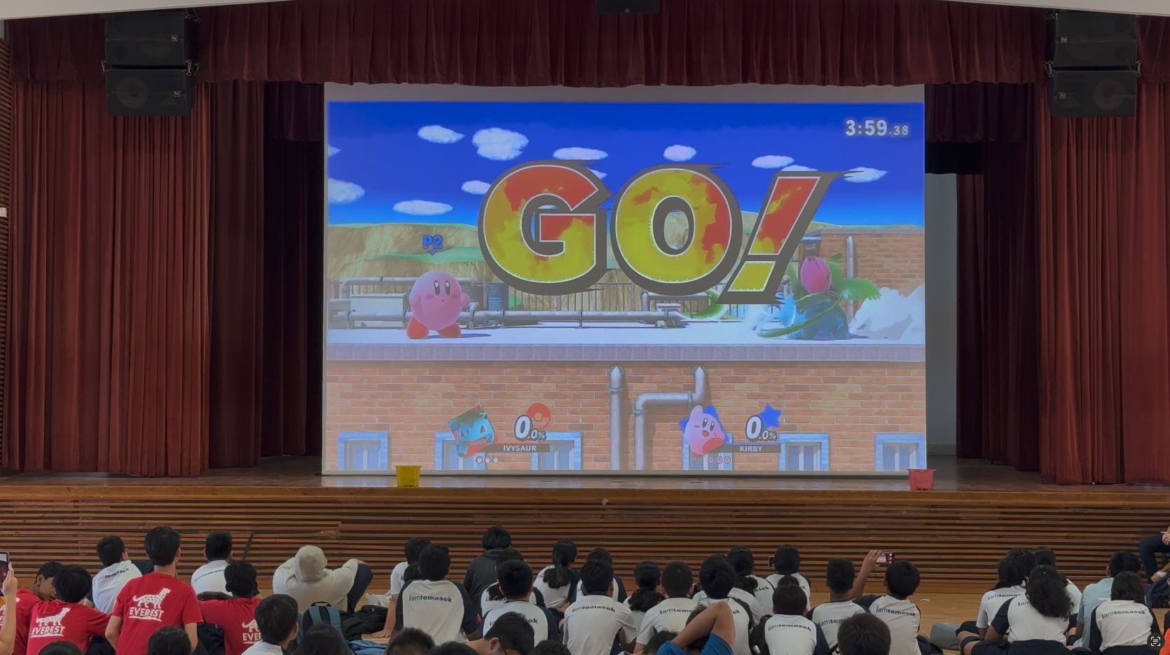 Students sit on a floor watching a large projected Super Smash Bros screen showing Kirby vs Ivysaur with GO! displayed.