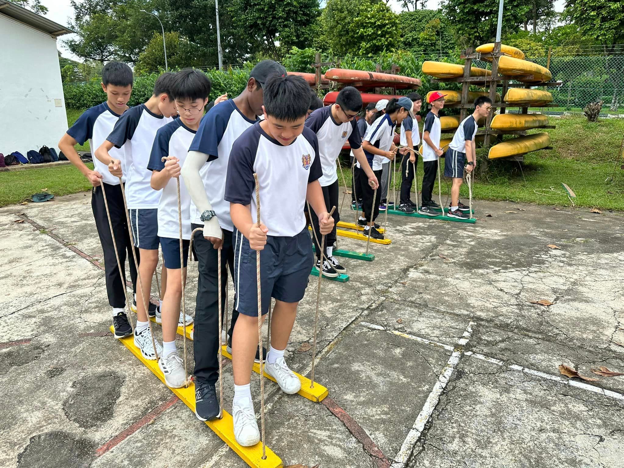 Students in uniforms balance on shared planks holding ropes during an outdoor team-building activity near stacked kayaks.