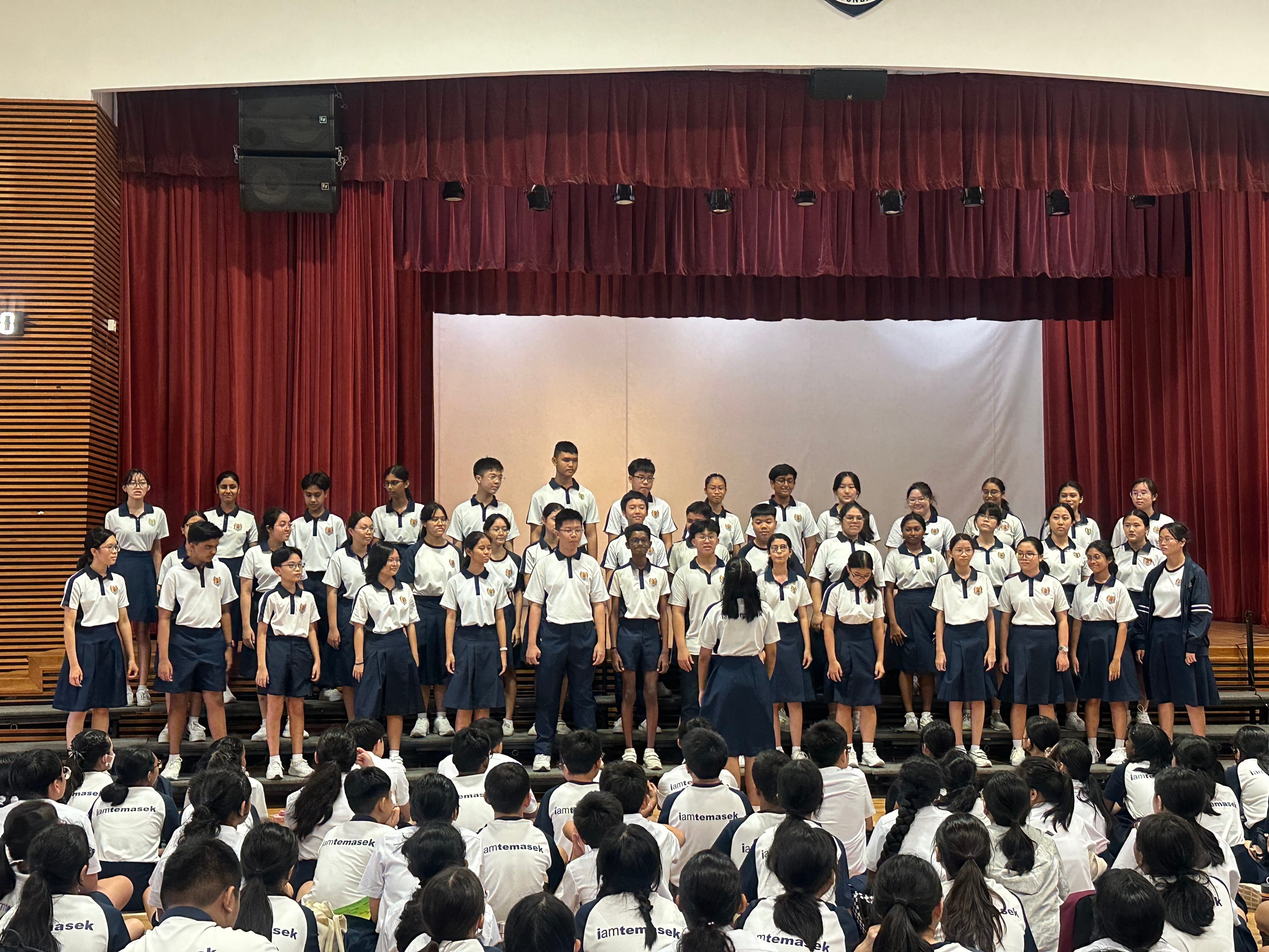 A school choir in white and navy uniforms performs on a stage with red curtains before a seated student audience.