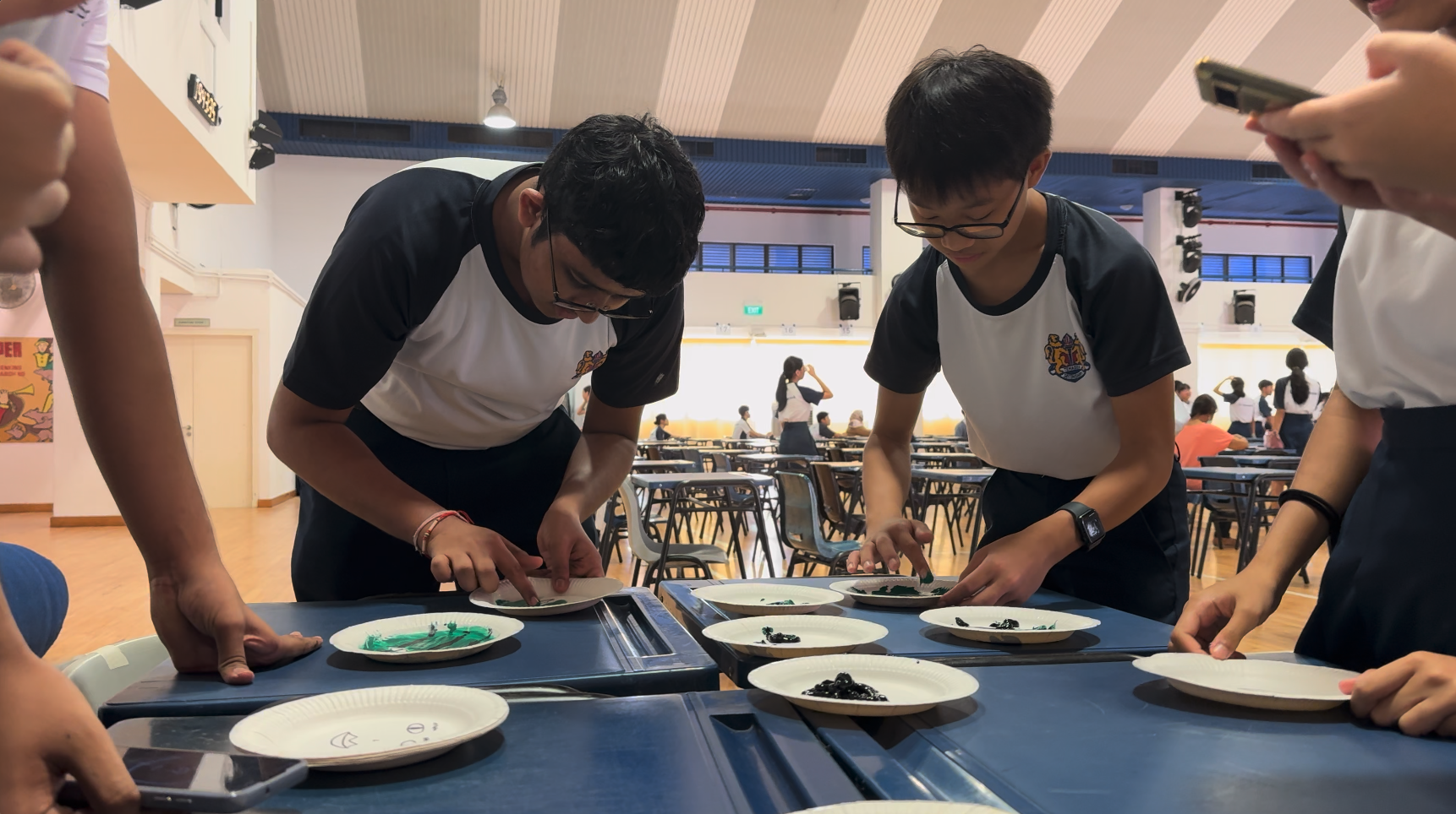 Two students in matching uniforms lean over a blue table with paper plates holding colorful materials in a school hall.