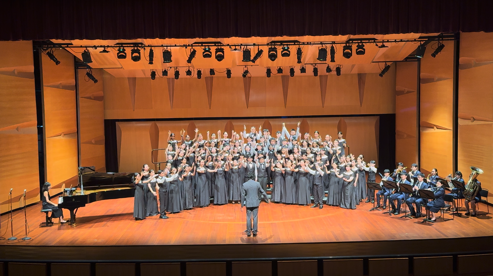 A conductor leads a large youth choir in gray formal wear on a concert hall stage with a pianist and brass ensemble.