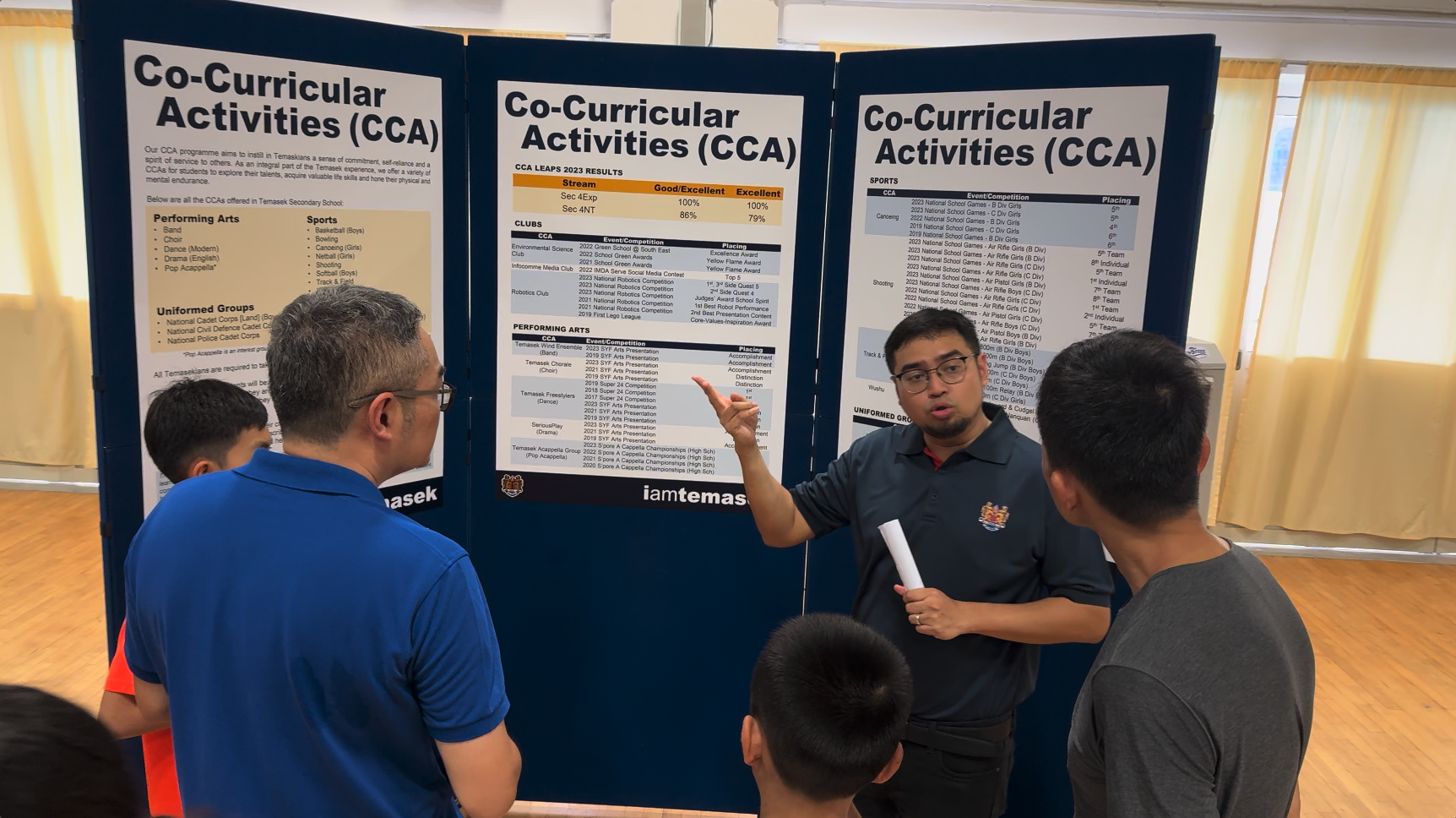 A man points at Co-Curricular Activities display boards while speaking to a small group of people.