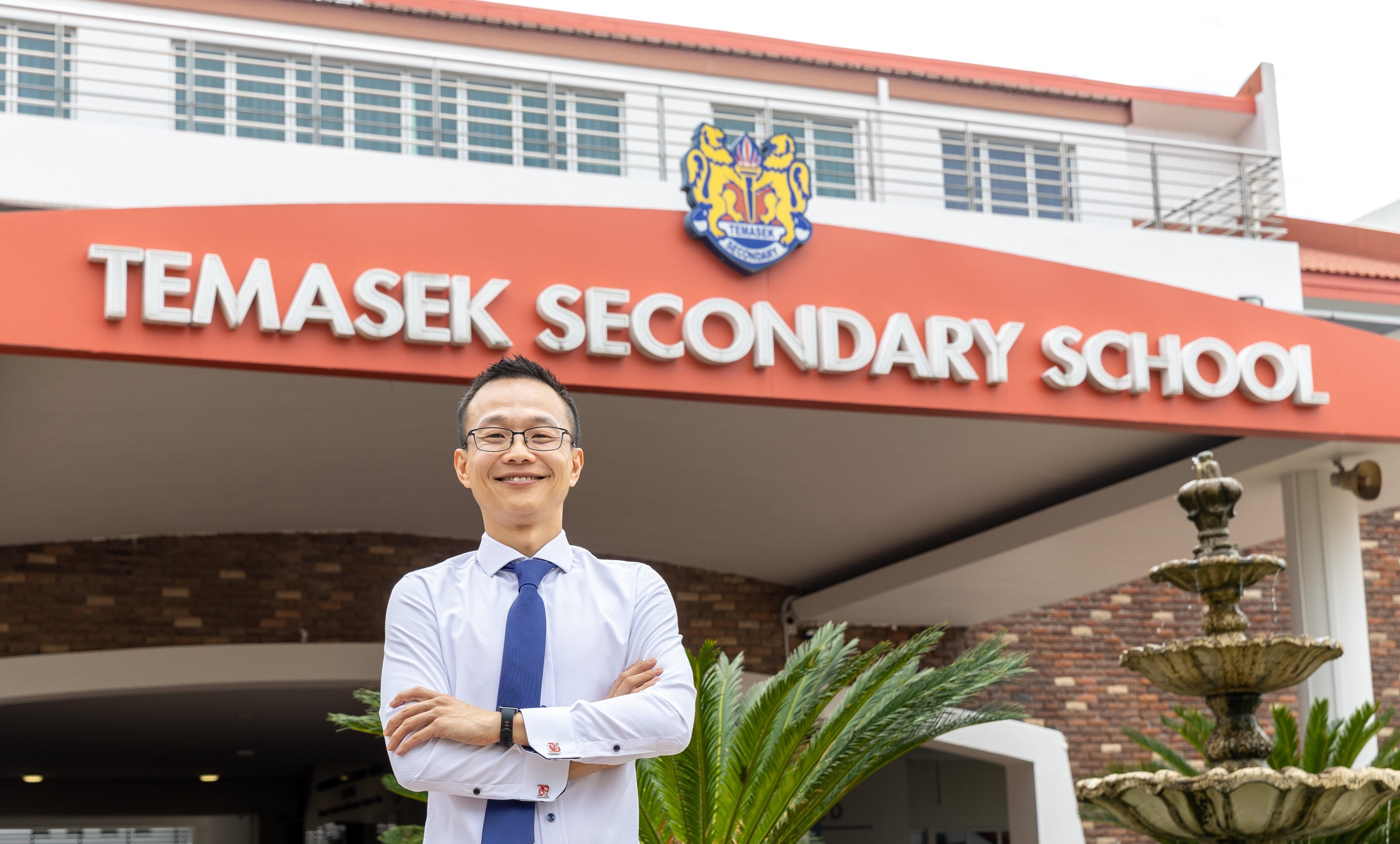 A smiling man in a white shirt and blue tie stands arms crossed before Temasek Secondary School's entrance sign.
