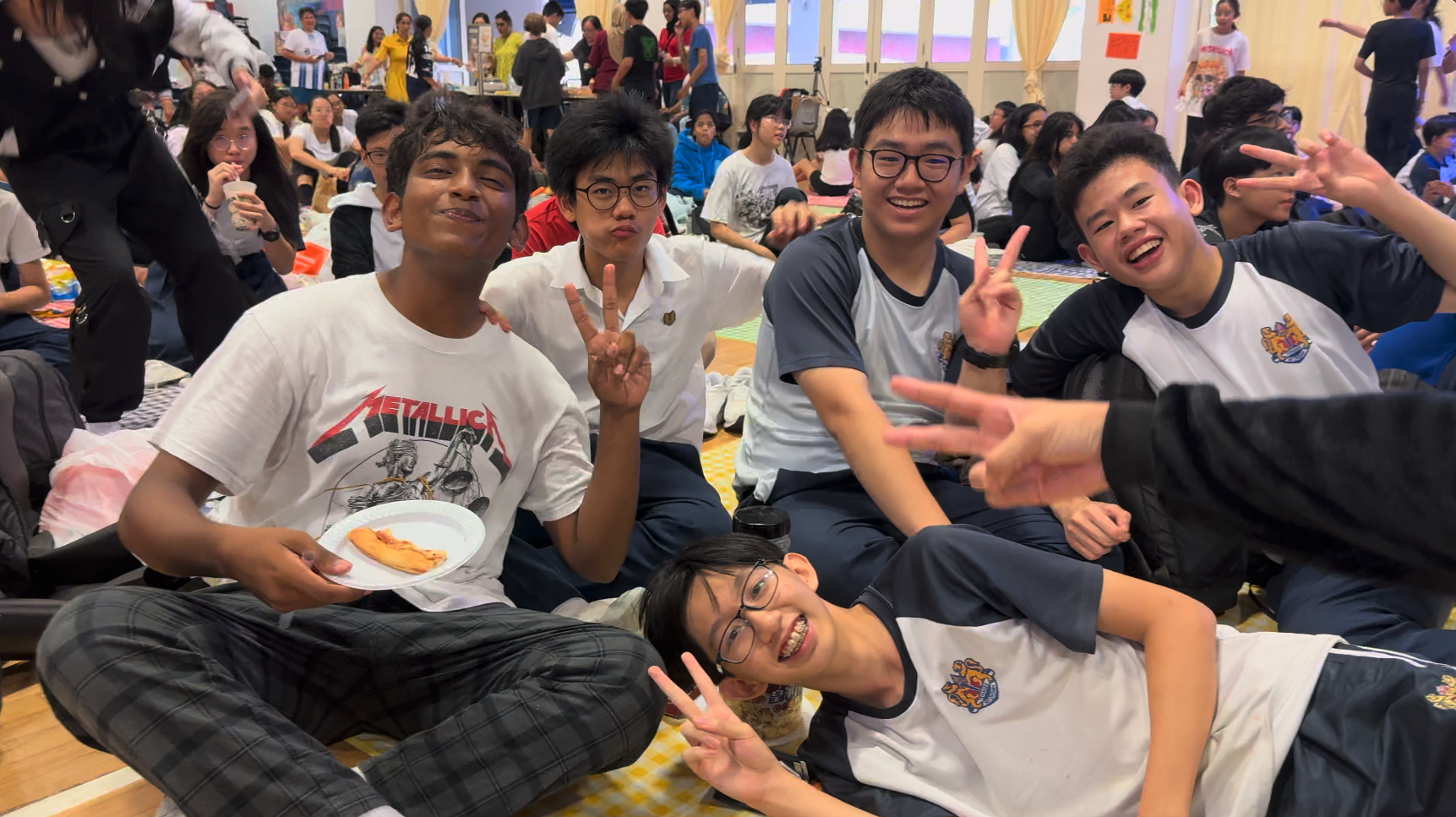 Five smiling teenage boys sit on a floor at a school event, posing with peace signs and holding a plate of food.