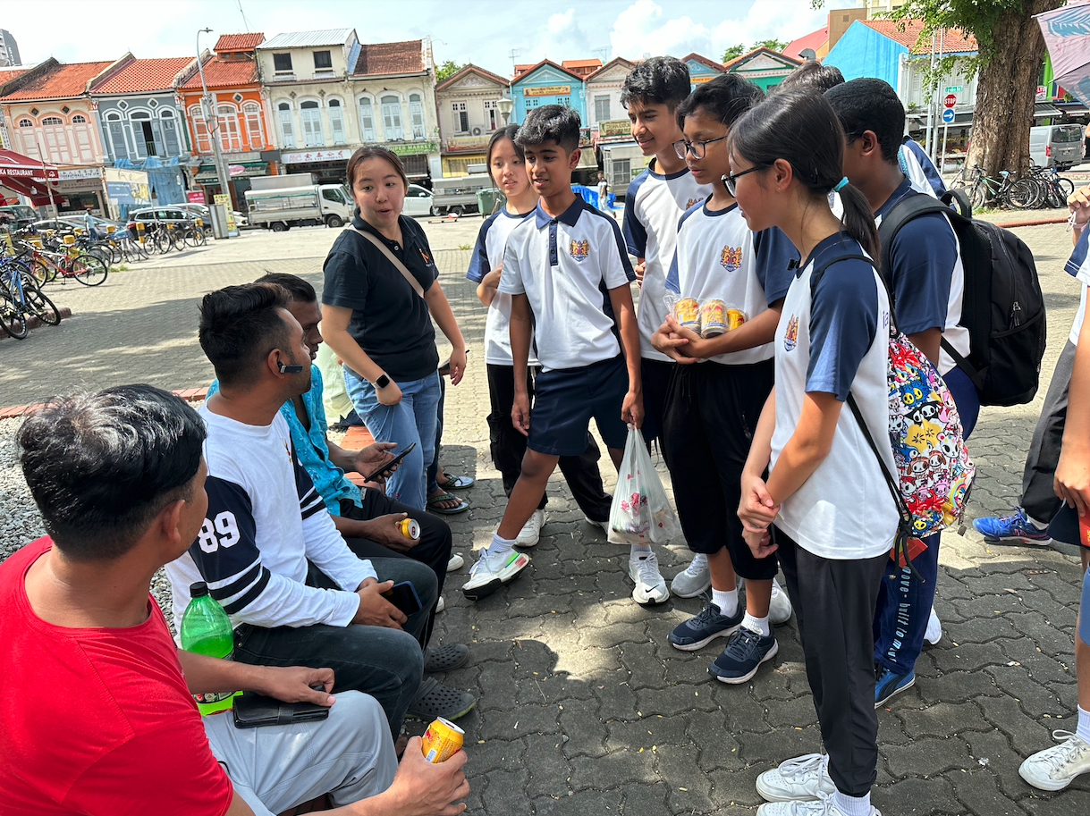 Students in white uniforms gather outdoors on a paved square, talking with adults, colorful shophouses visible behind them.