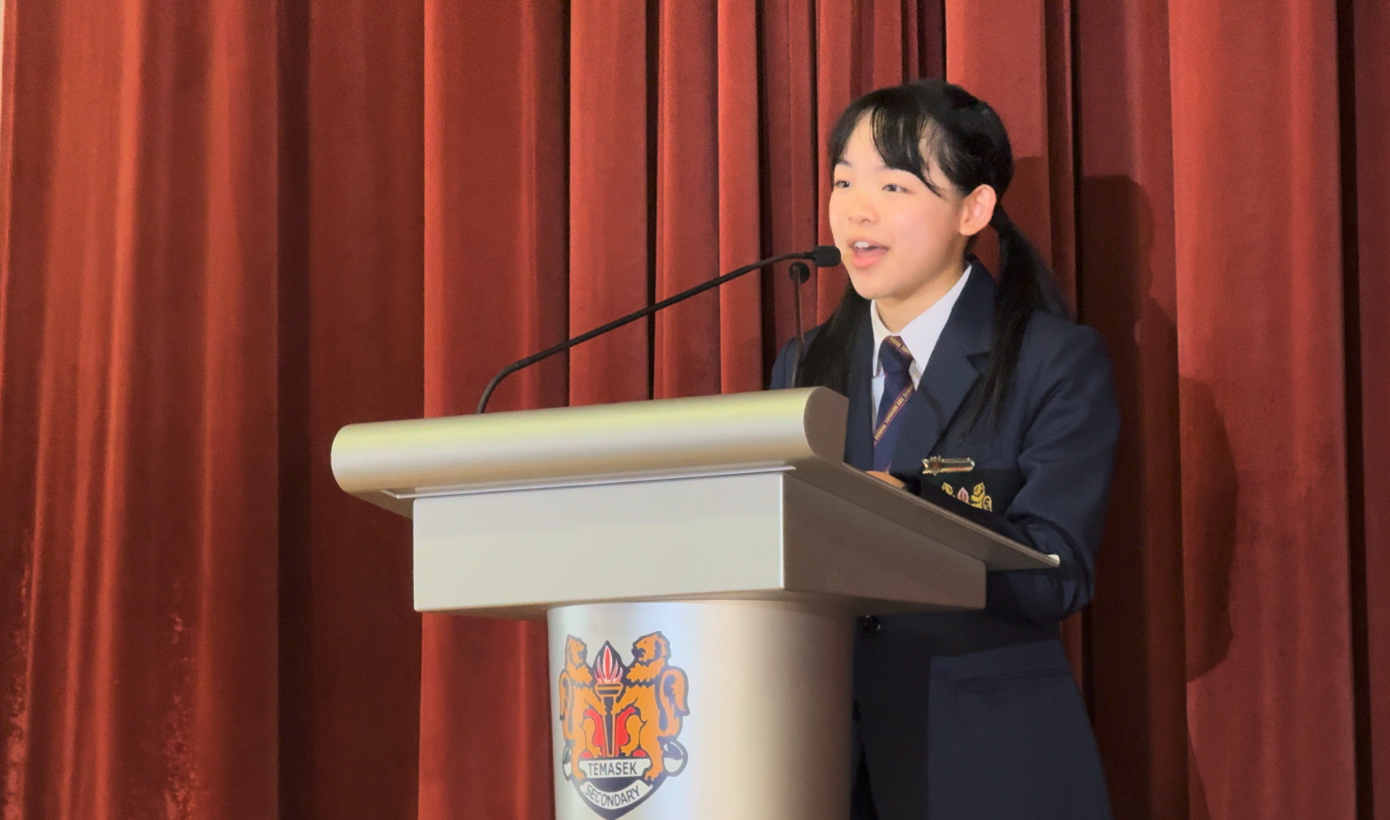 A young girl in a navy school uniform speaks at a Temasek Secondary podium before red curtains.