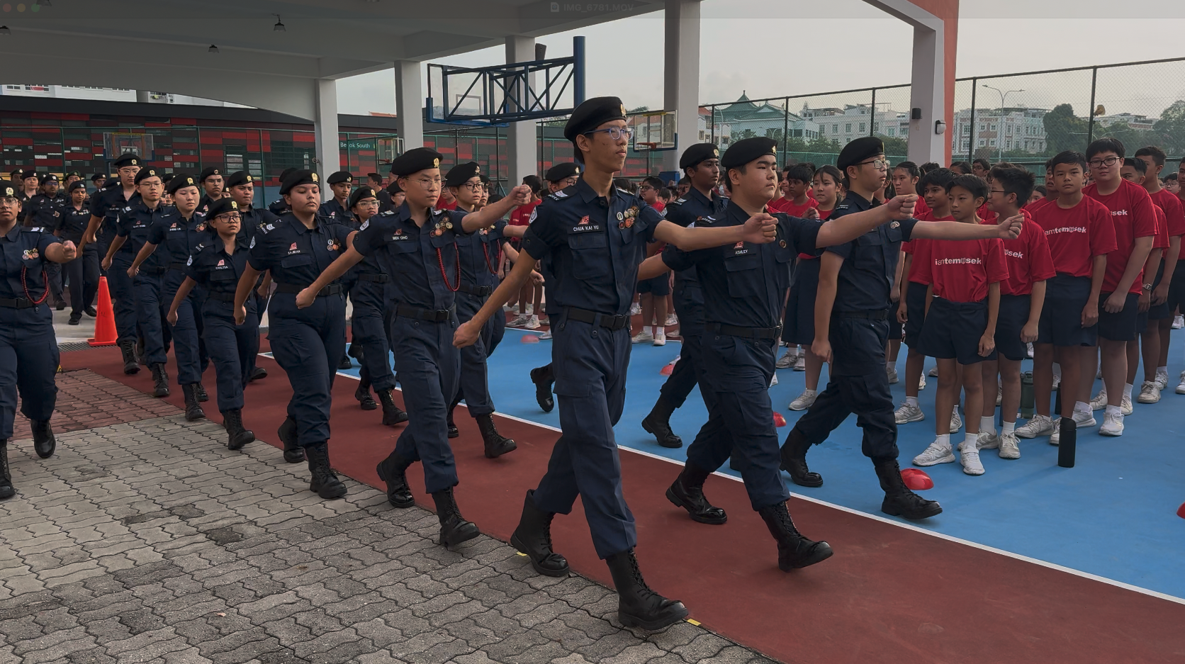 Students in navy uniforms and black berets march in formation past spectators in red shirts at a school courtyard.