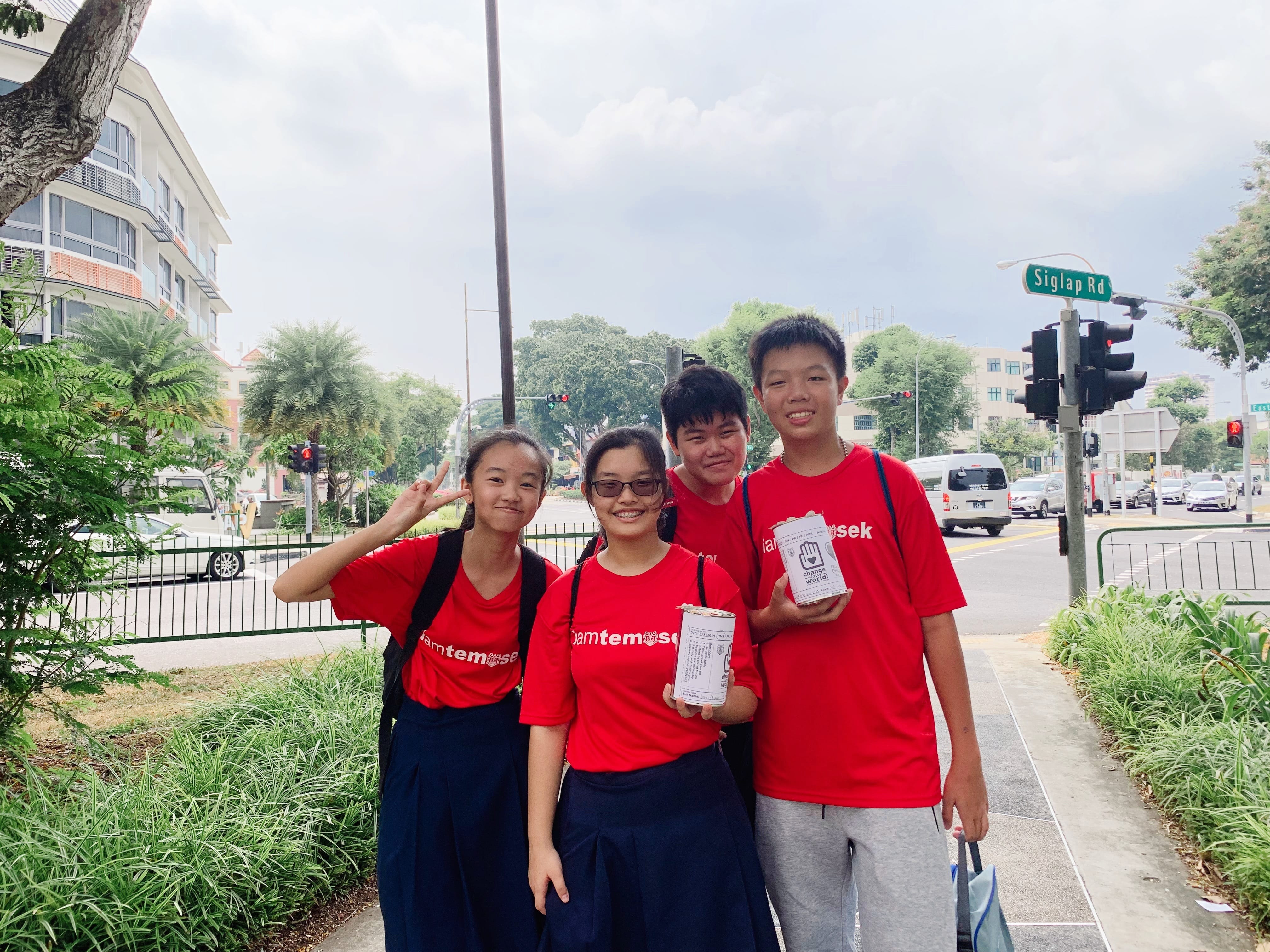 Four students in red Temasek shirts smile on a Singapore sidewalk near Siglap Rd, holding donation cans.
