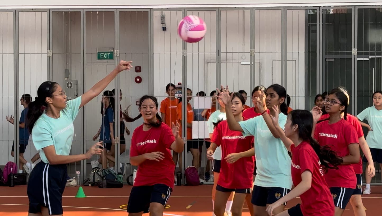 Girls in red and mint green shirts play volleyball indoors, with one player reaching up to hit a pink ball.