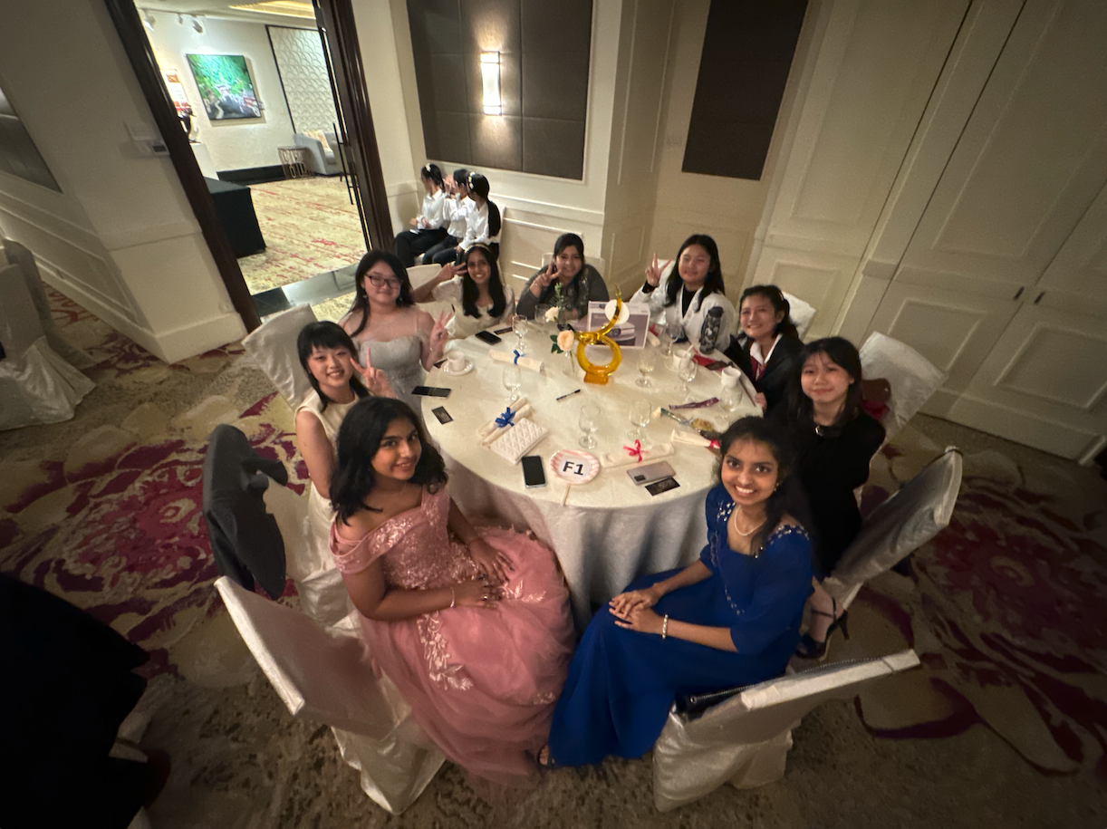 A group of young women in formal dresses sit around a decorated banquet table, smiling and posing for the camera.