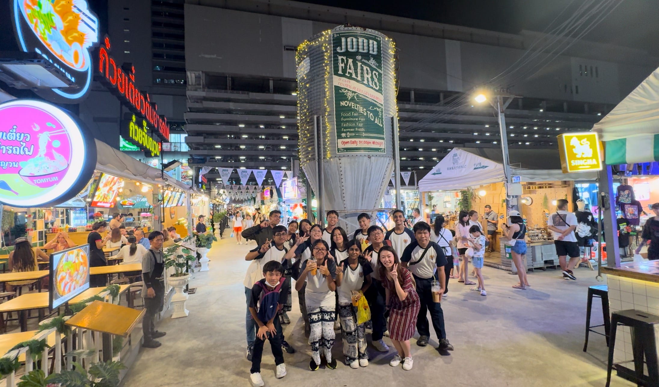 A group of people pose at Jodd Fairs night market in Bangkok, surrounded by lit food stalls and Thai signage.