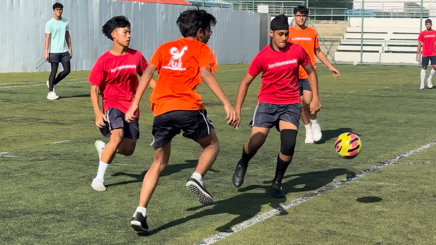 Young players in red and orange shirts compete for a soccer ball on a green turf field.