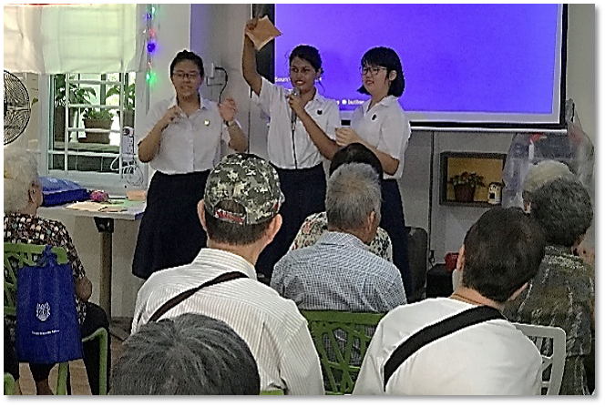 Three students in white uniforms present at the front of a room to a seated elderly audience near a purple screen.