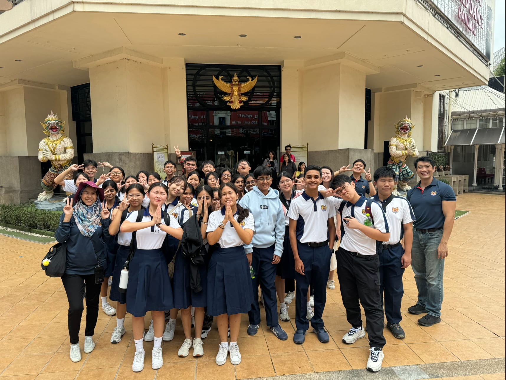 A large group of students in school uniforms poses outside a Thai building with golden Garuda and guardian statues.