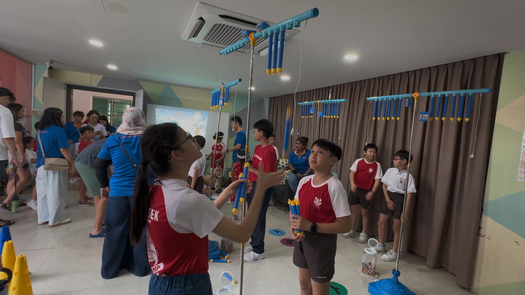 Children in red and white uniforms build tall blue tube structures on poles inside a colorful indoor room.