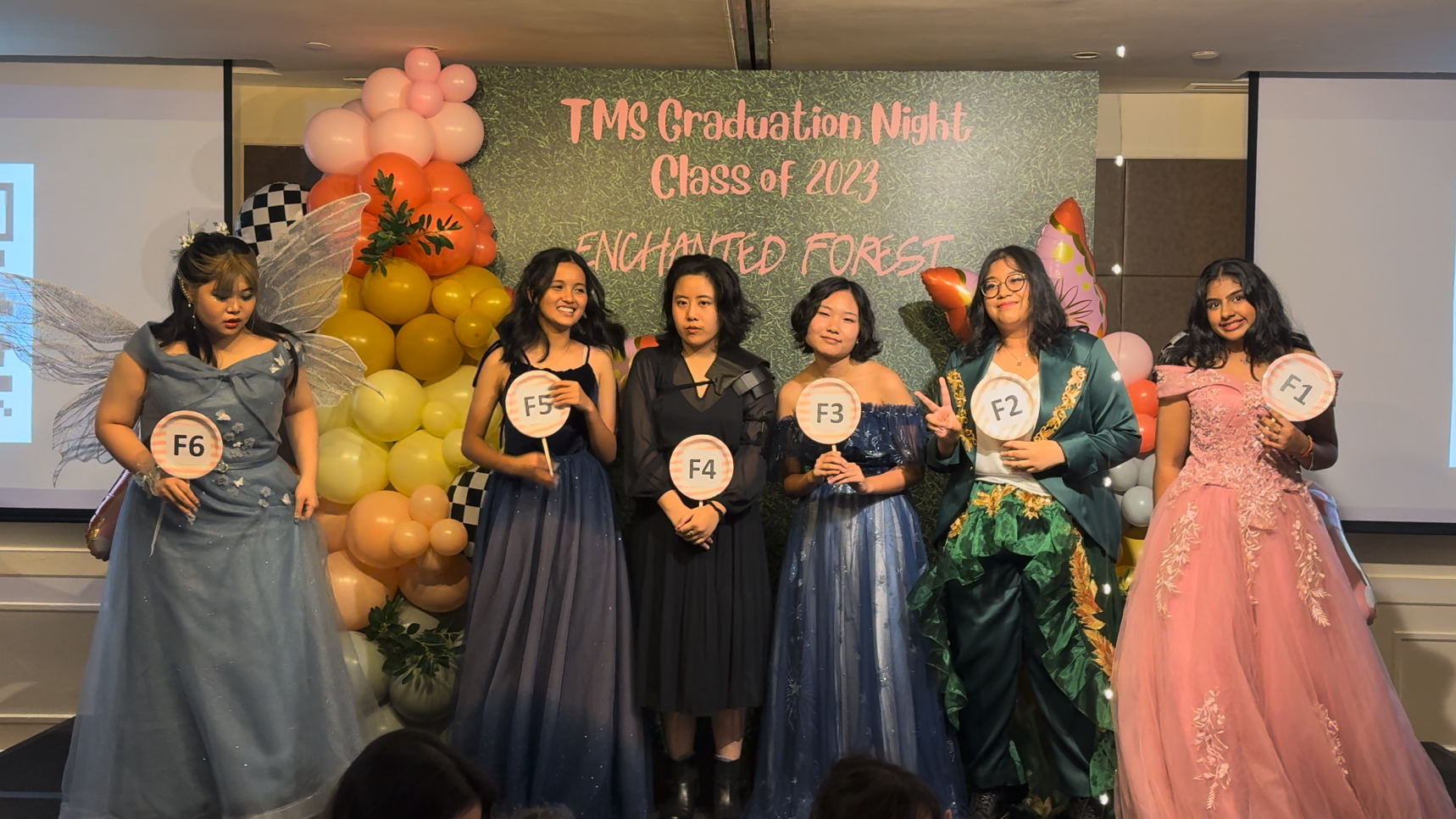 Six young women in formal gowns hold numbered paddles at TMS Graduation Night Class of 2023 Enchanted Forest event.