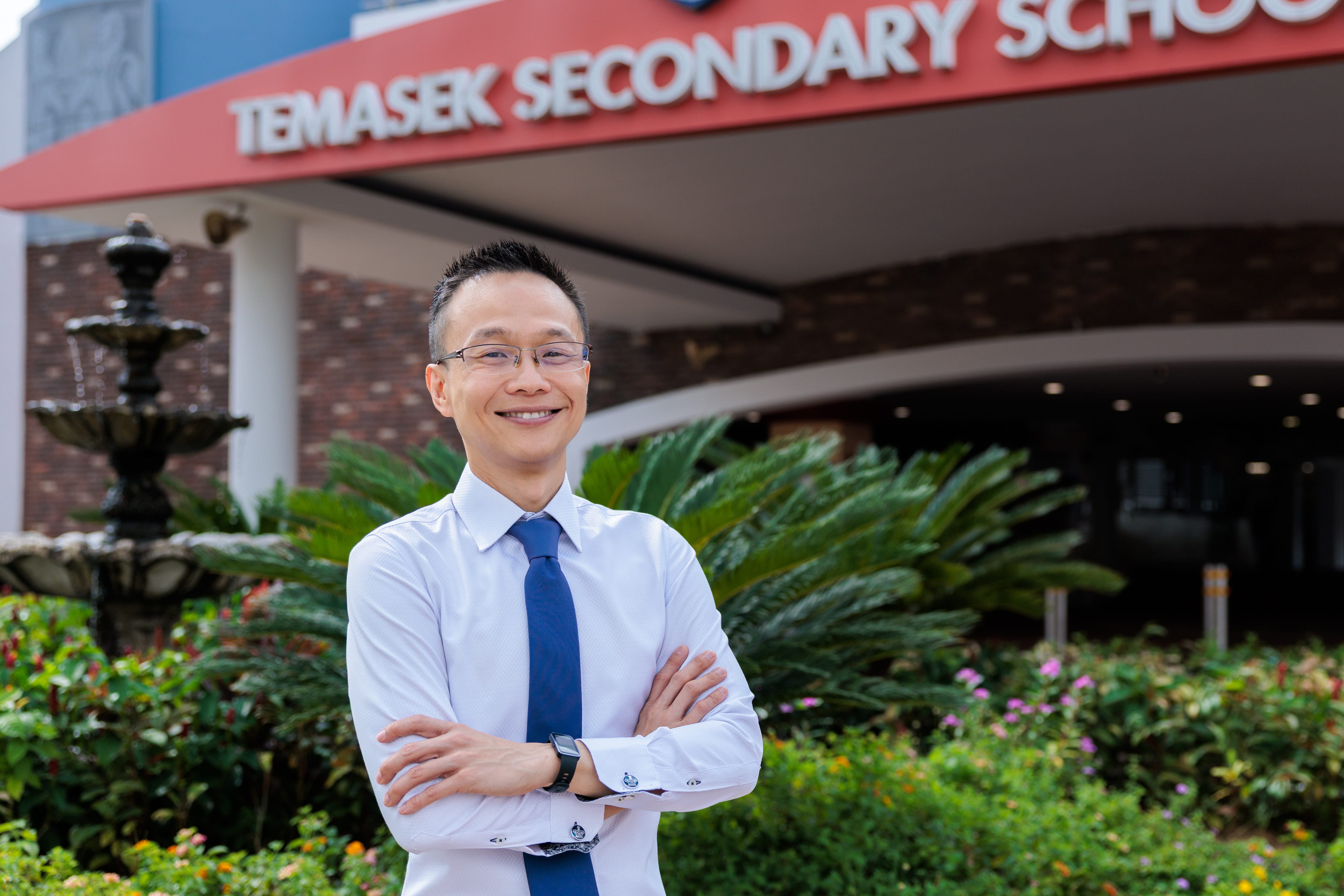 Smiling man in glasses with arms crossed stands outside Temasek Secondary School entrance and garden.