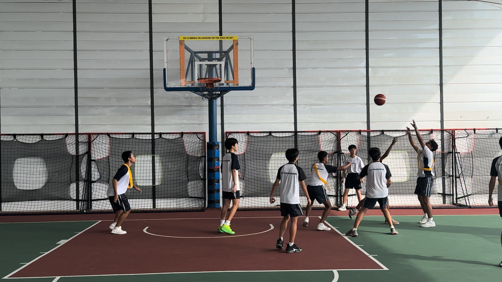 Several young male students in white uniforms play basketball on an indoor court, with one player shooting toward the hoop.