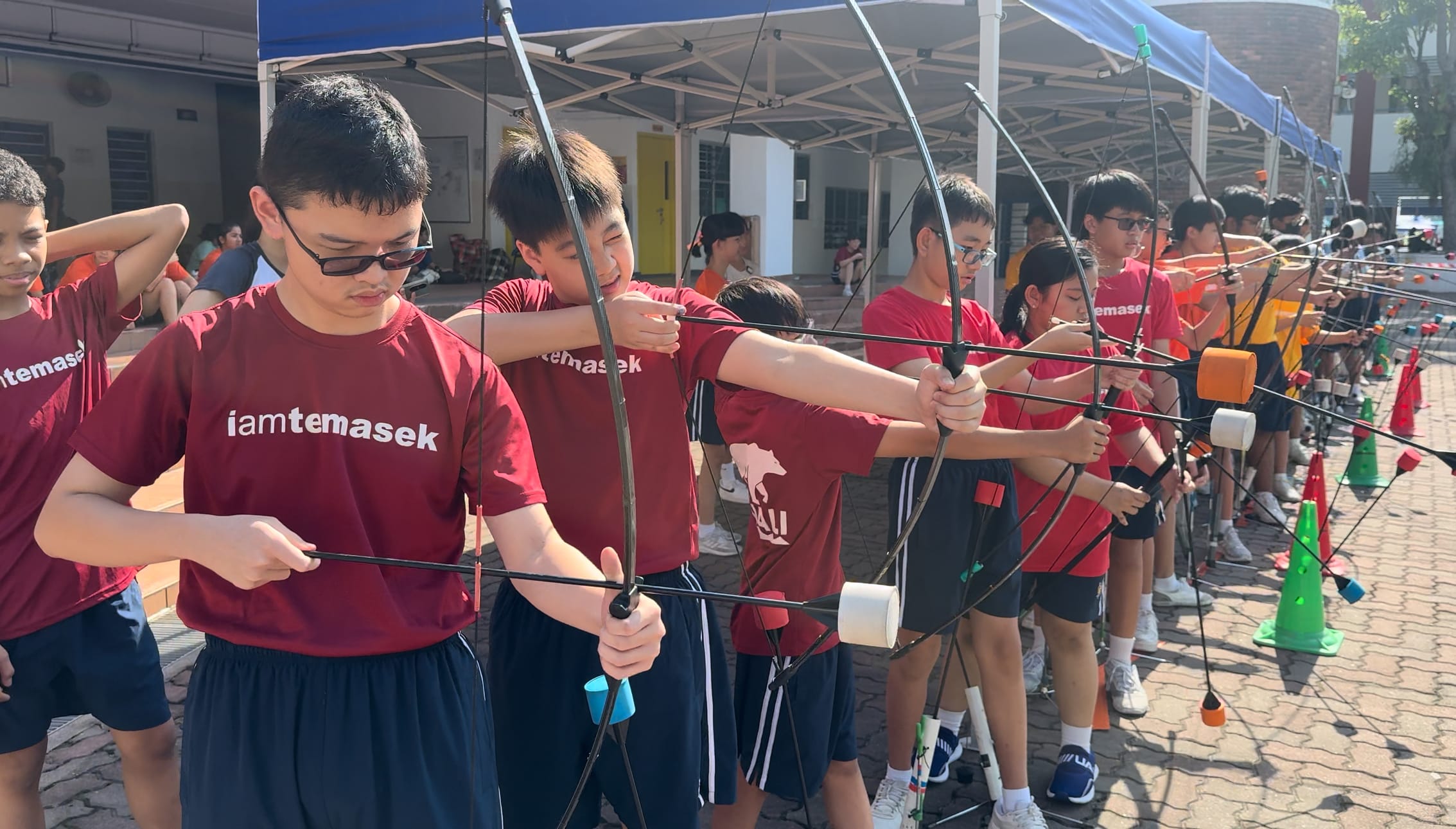 Students in red "iamtemasek" shirts stand in a line practicing archery with bows outdoors under blue canopies.