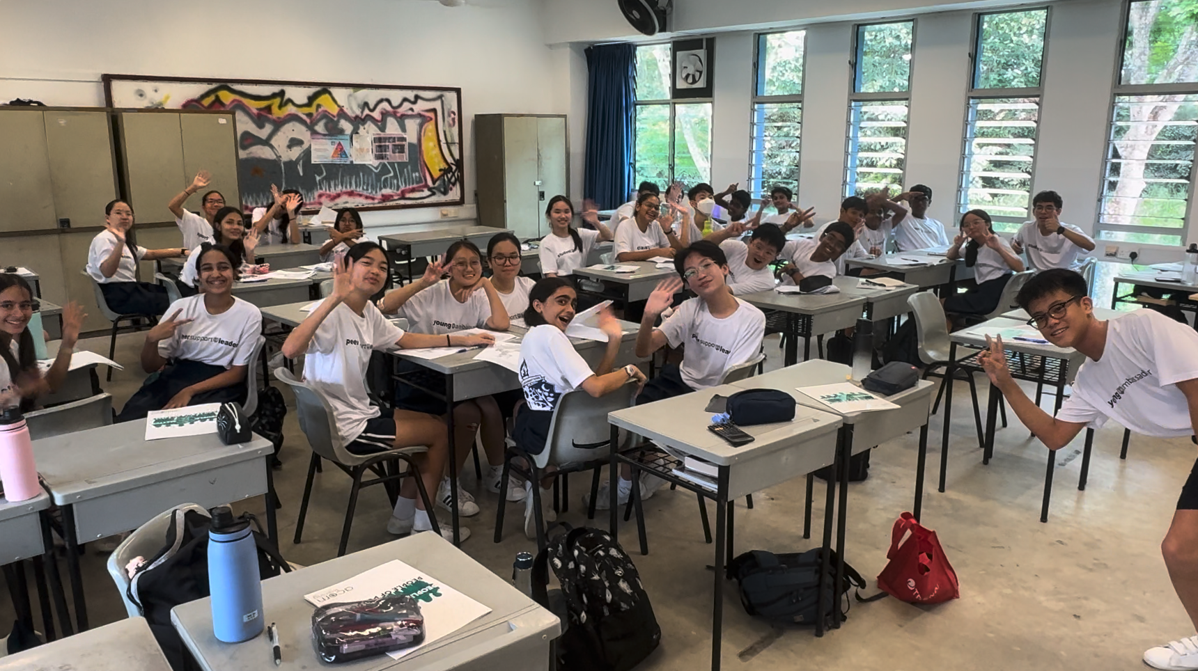A large group of students in white t-shirts sitting at desks in a classroom, smiling and waving at the camera.