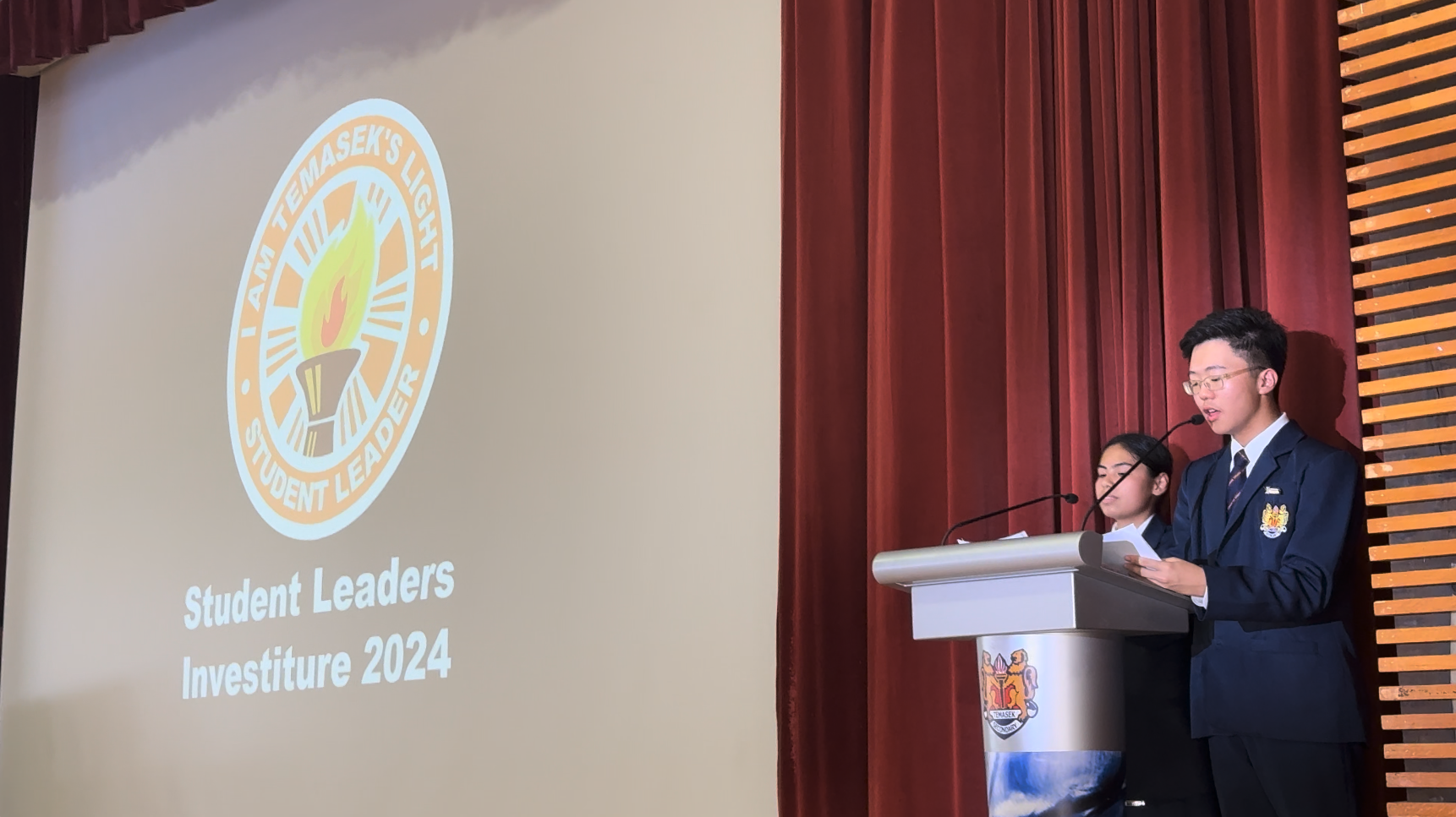 Two students in school uniforms speak at a podium beside a projected slide reading Student Leaders Investiture 2024.