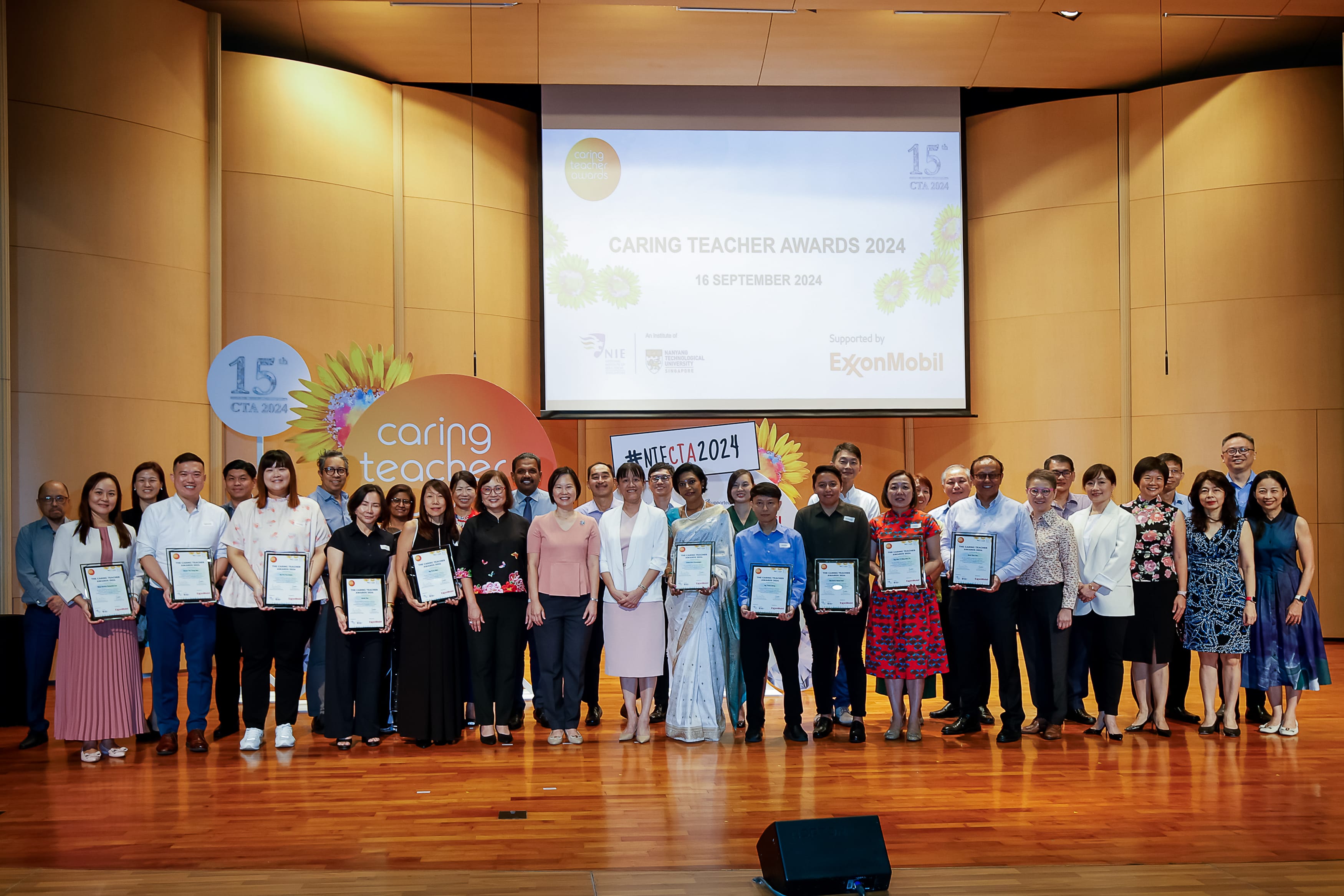 Group on stage holding certificates under Caring Teacher Awards 2024 screen and large caring teacher backdrop
