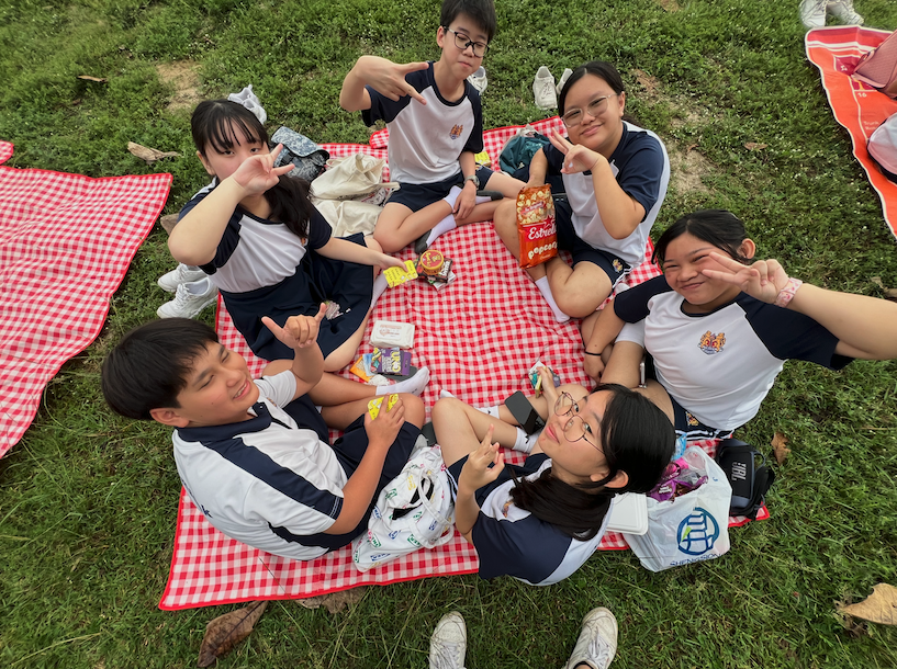 Six students in matching uniforms sit on a red checkered picnic blanket on grass, posing with peace signs and snacks.
