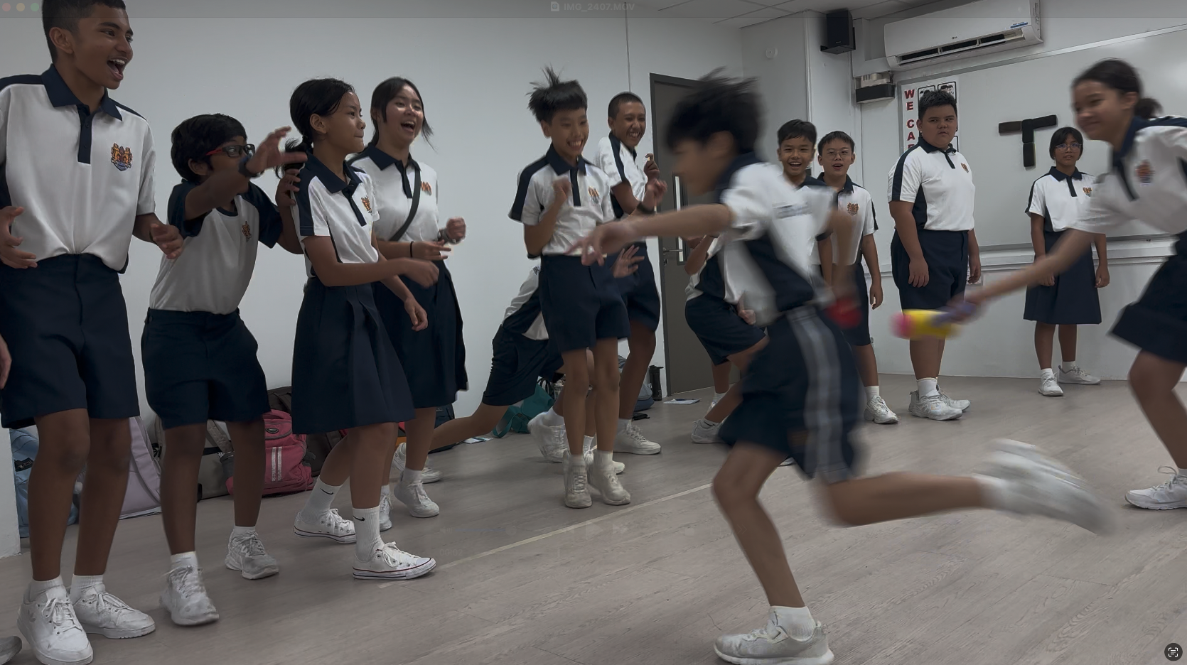 Students in white and navy uniforms laugh and cheer as one runs past holding a colorful object indoors.