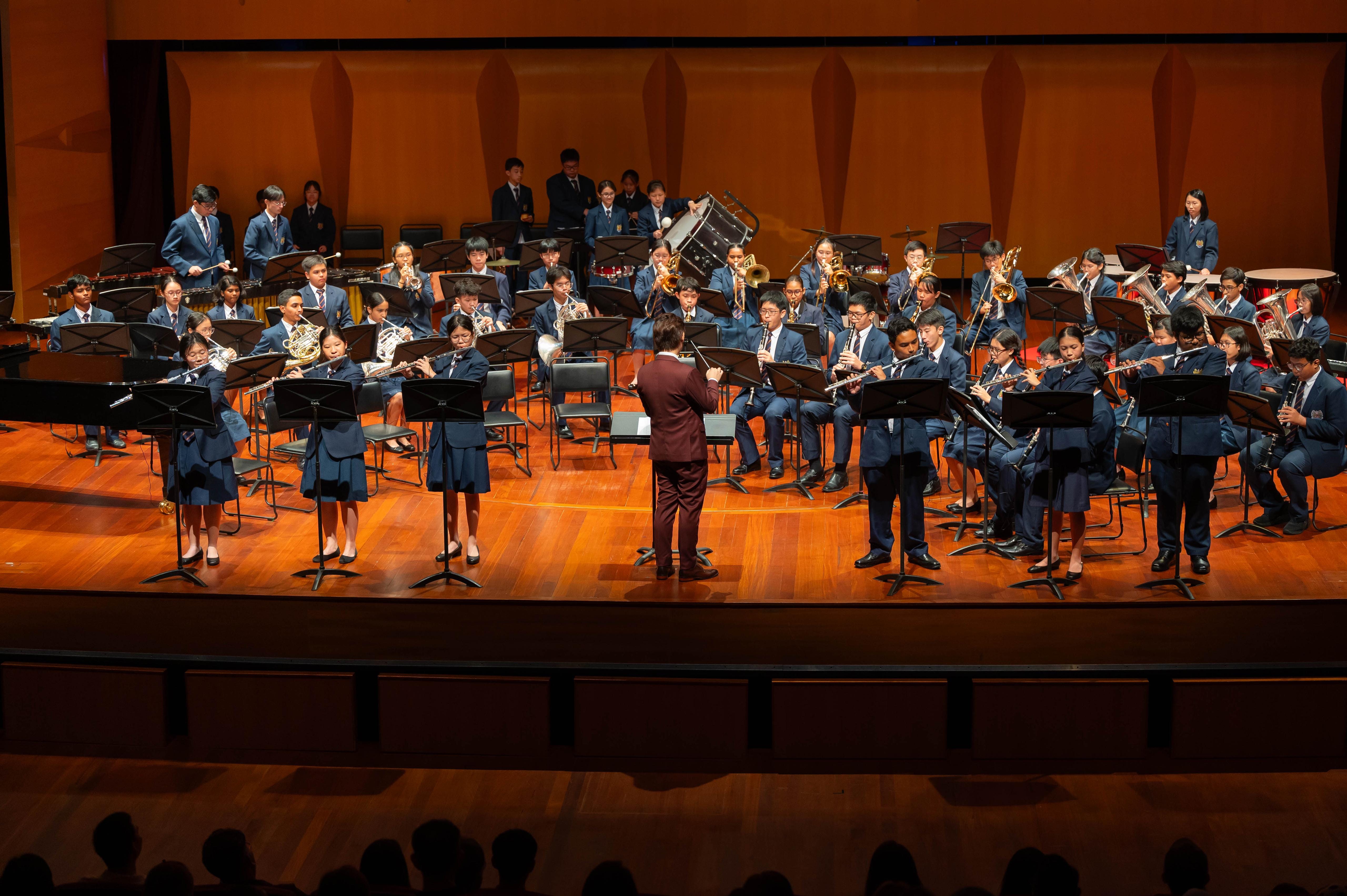 A conductor in burgundy leads a large youth wind band in blue uniforms performing on a wooden concert stage.