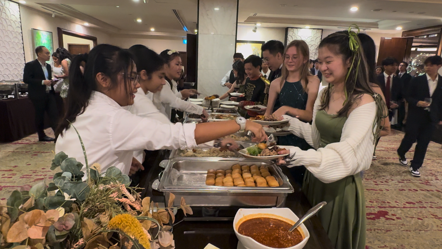 Formally dressed guests being served food at a buffet line by staff in white shirts at an indoor banquet event.