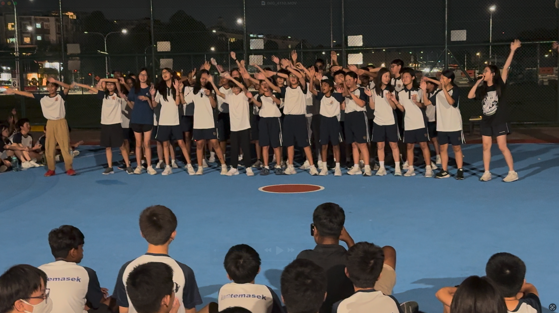 A large group of students in white and navy uniforms raise their hands on an outdoor court at night before a seated audience.