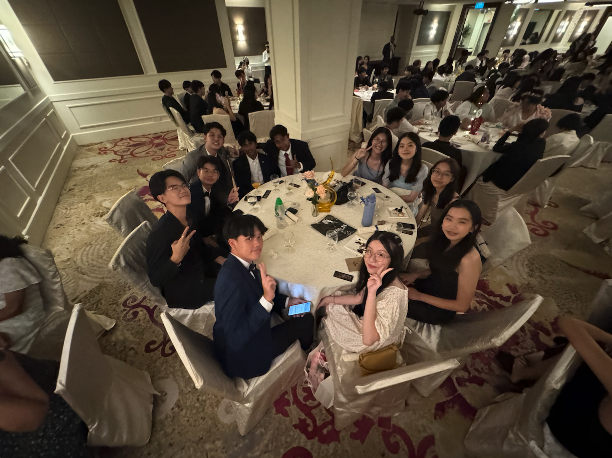 Young people in formal attire sit around a round table in a crowded banquet hall, smiling and posing for the camera.