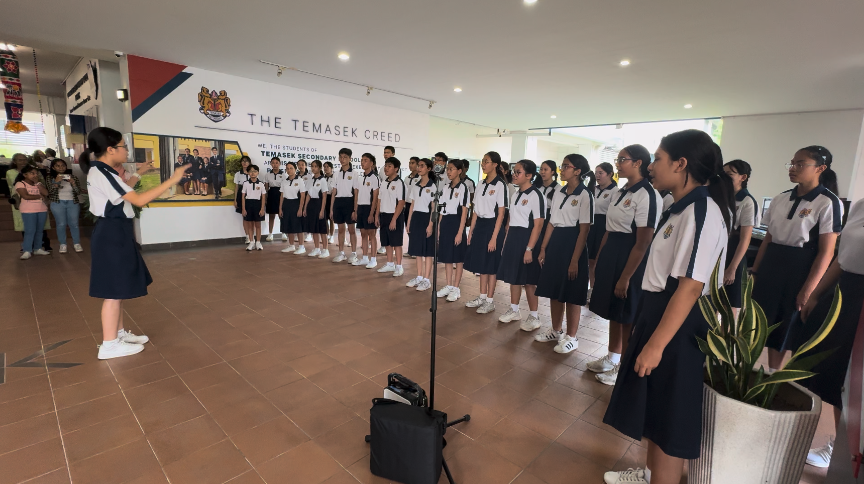 Students in white and navy uniforms perform as a choir at Temasek Secondary School, led by a conductor.