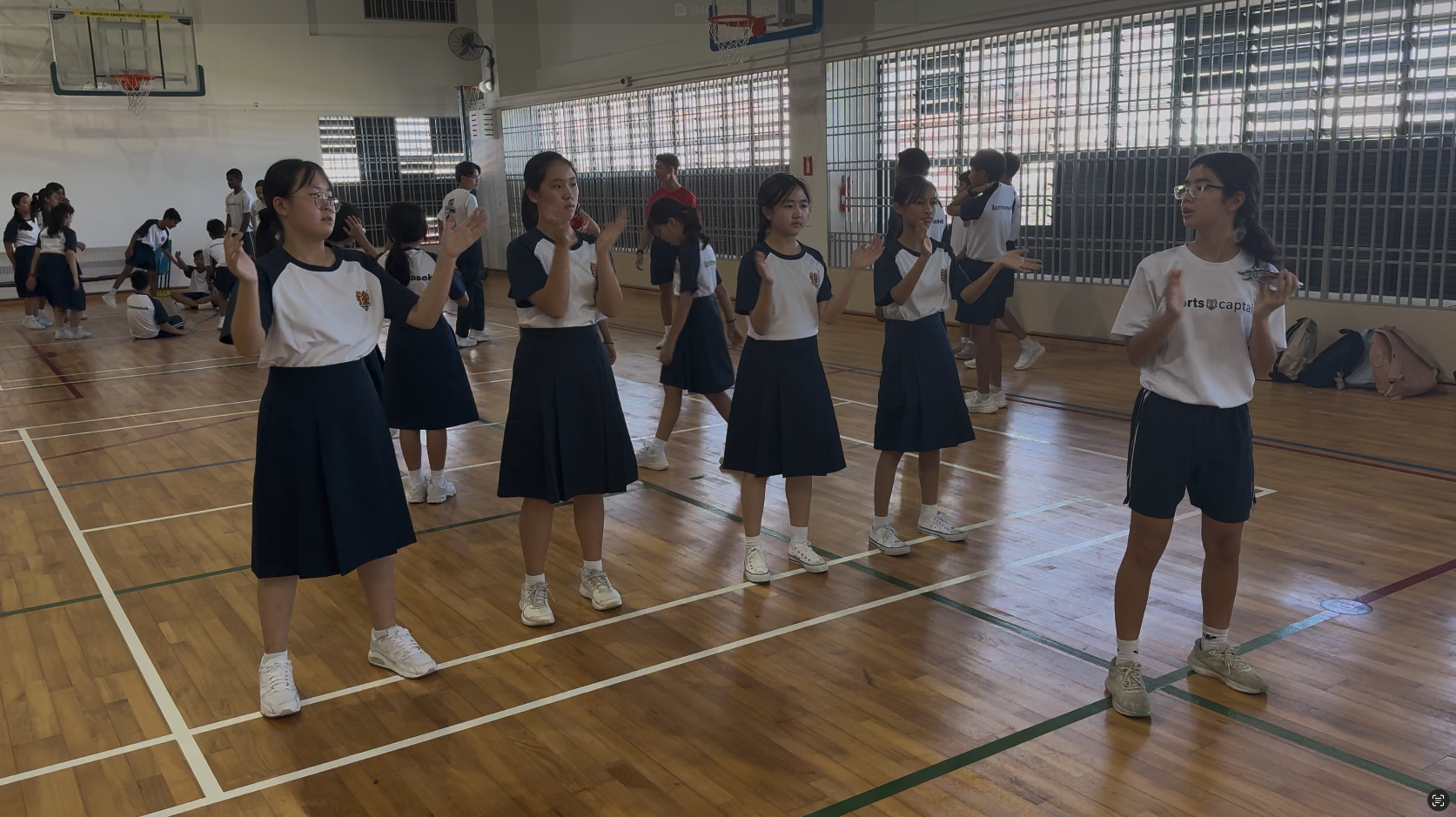 Students in school uniforms perform a dance or exercise routine inside a gymnasium with wooden floors.