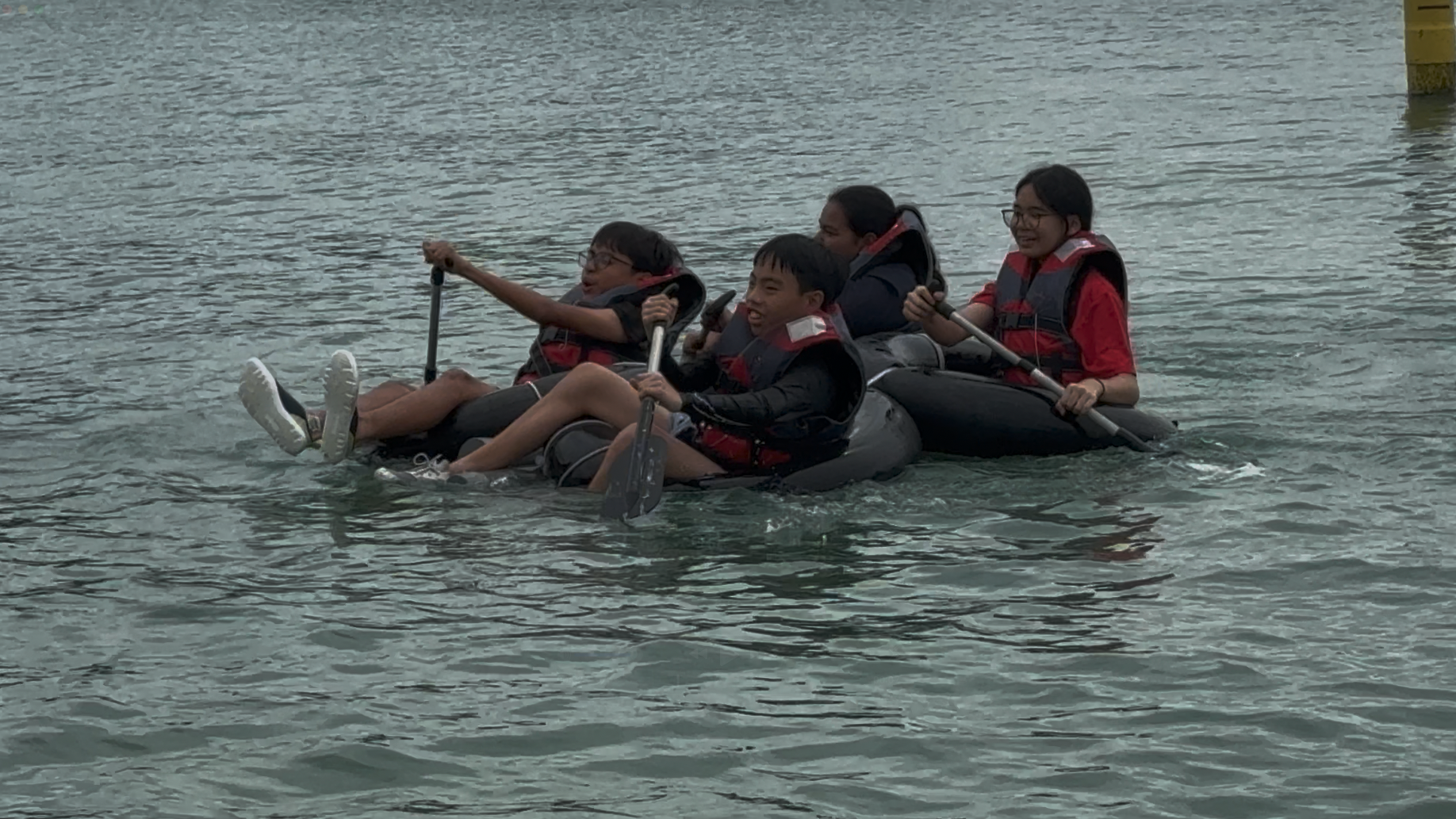Four children wearing red and black life jackets paddle together on inflatable tubes in open water.
