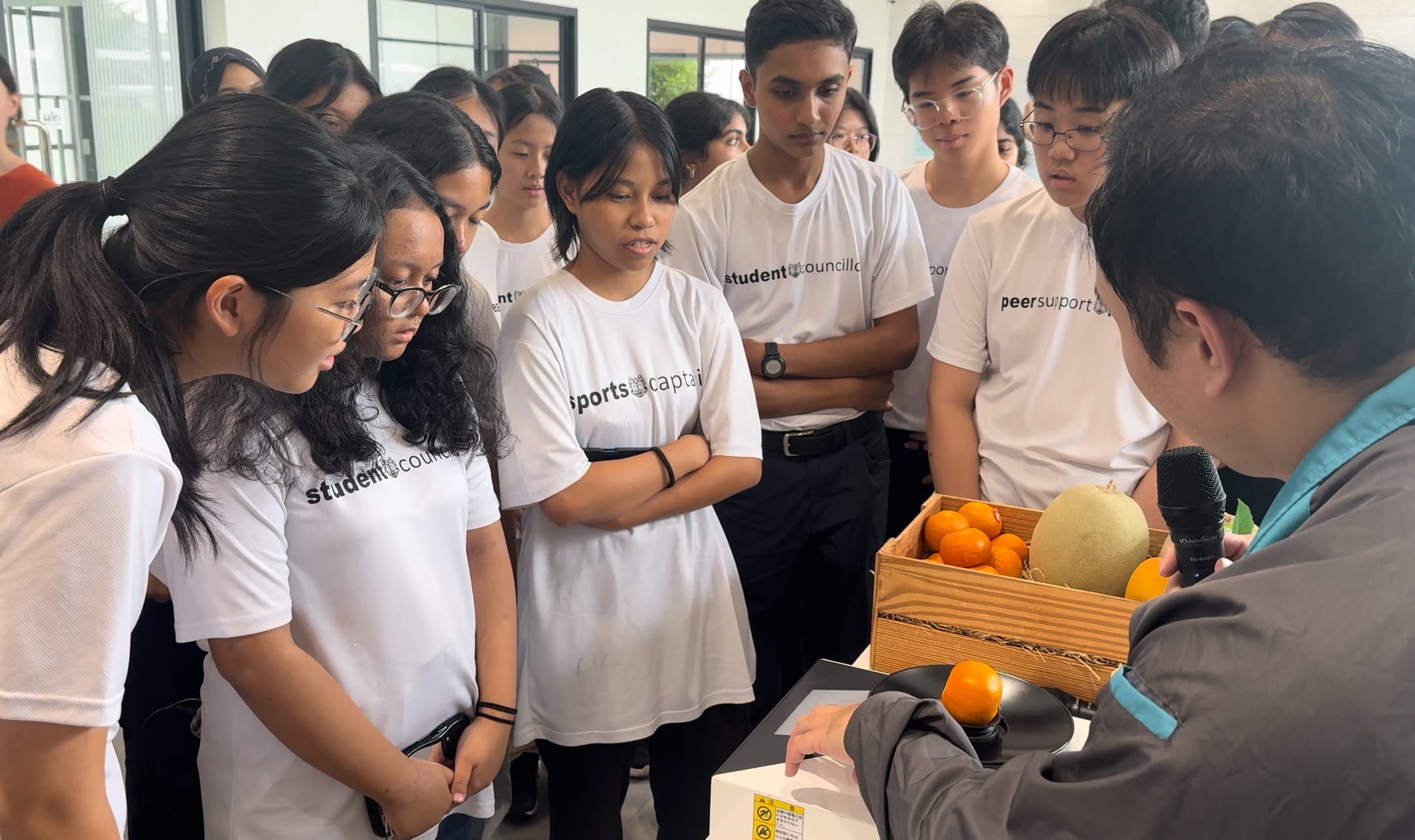Students in white council and sports captain shirts gather around a presenter holding a mic near a crate of oranges and a melon.