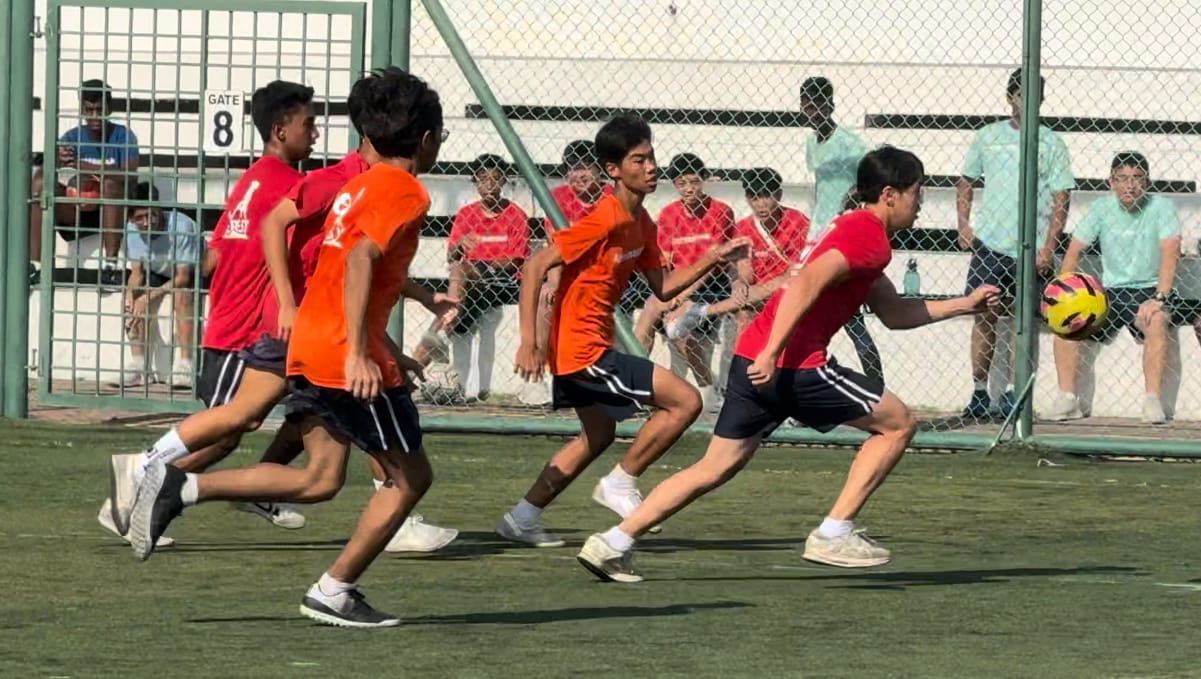 Young boys in red and orange shirts play soccer on a green field with spectators watching behind a fence.