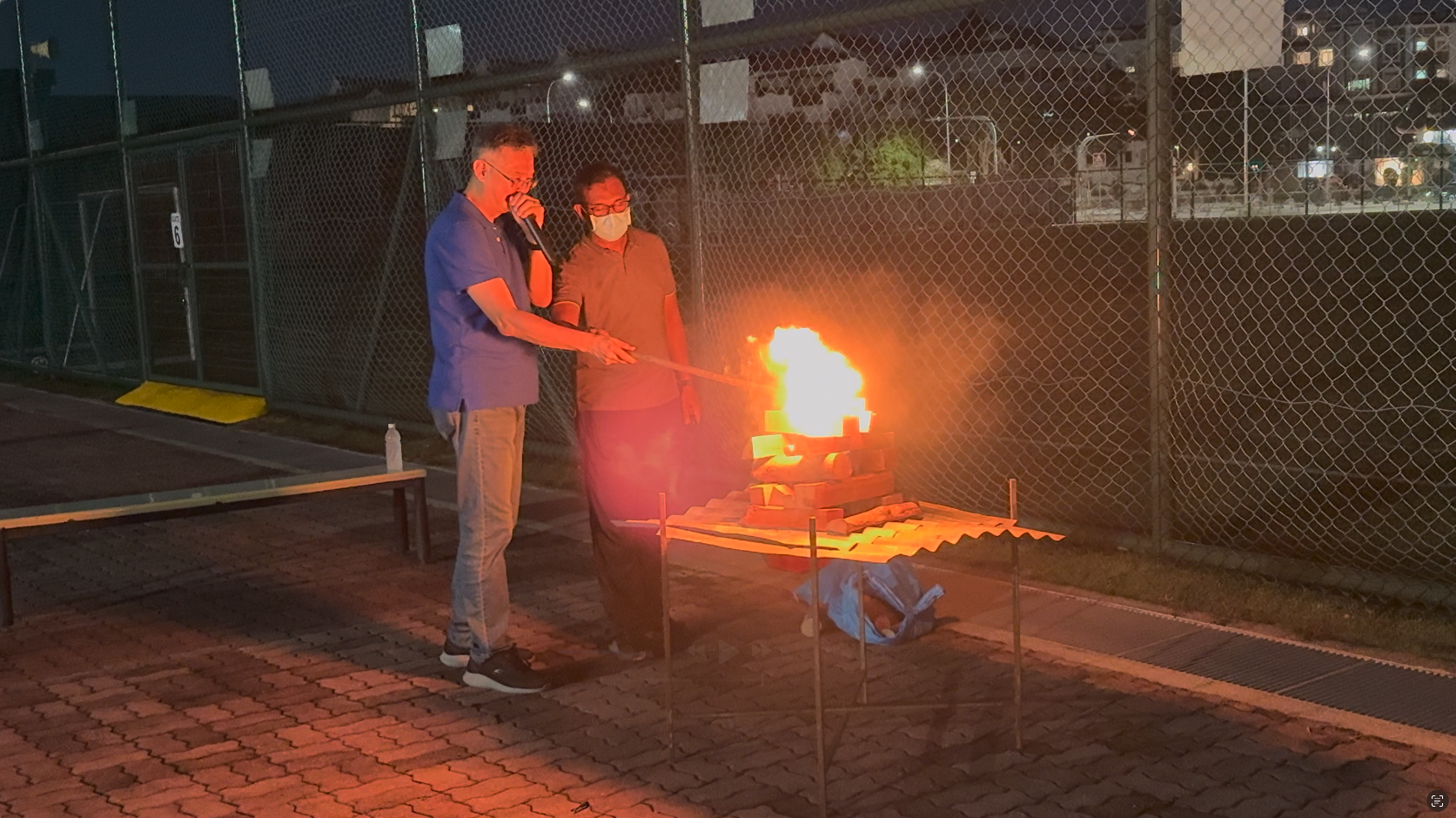 Two masked men stand outdoors at night near a metal rack with a large open flame burning on it.