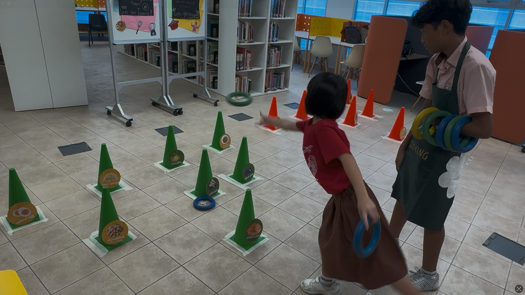 Two children playing a ring toss game with green and orange cones on a library floor.