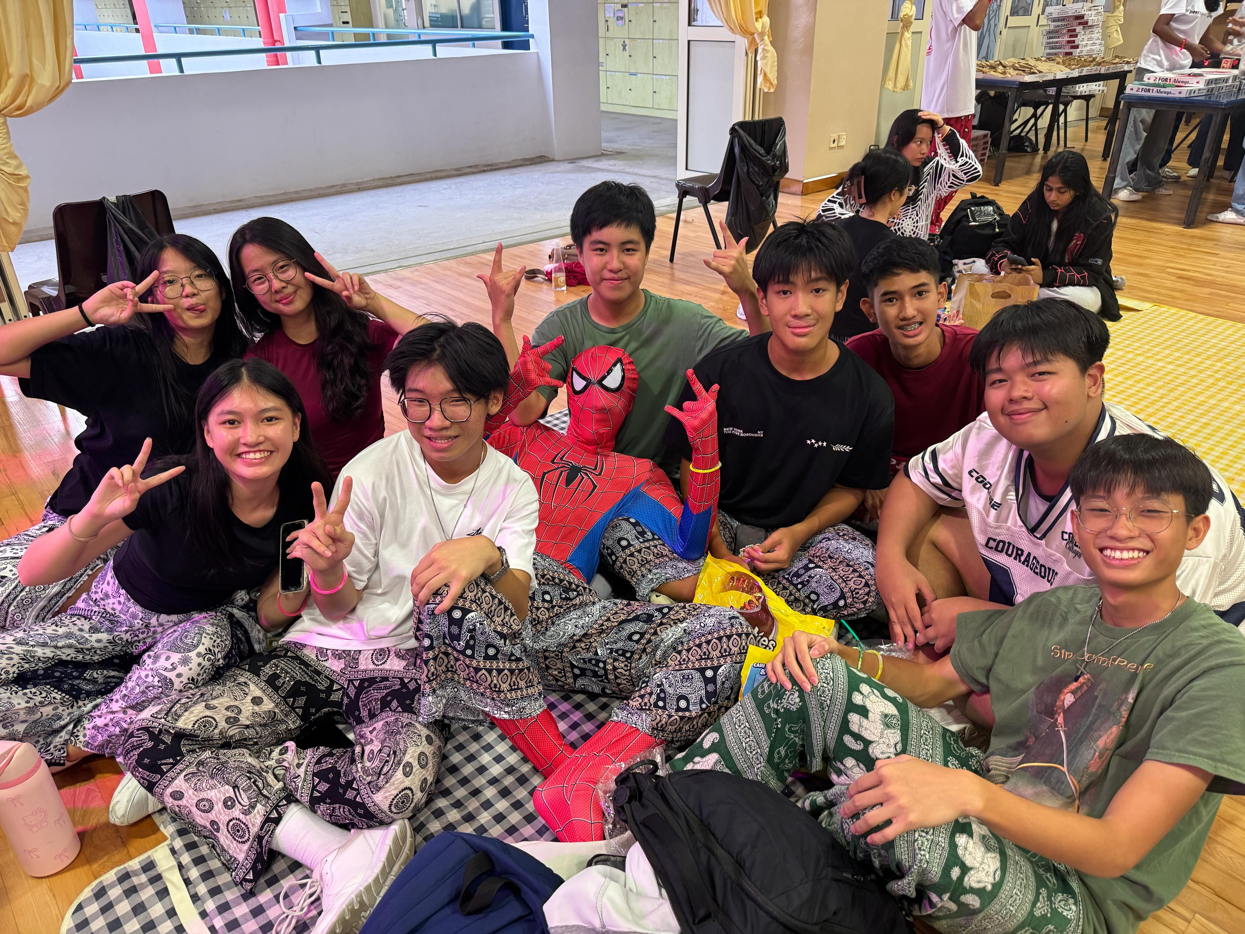 A group of teenagers sitting on a floor posing with peace signs, one wearing a full Spider-Man costume.