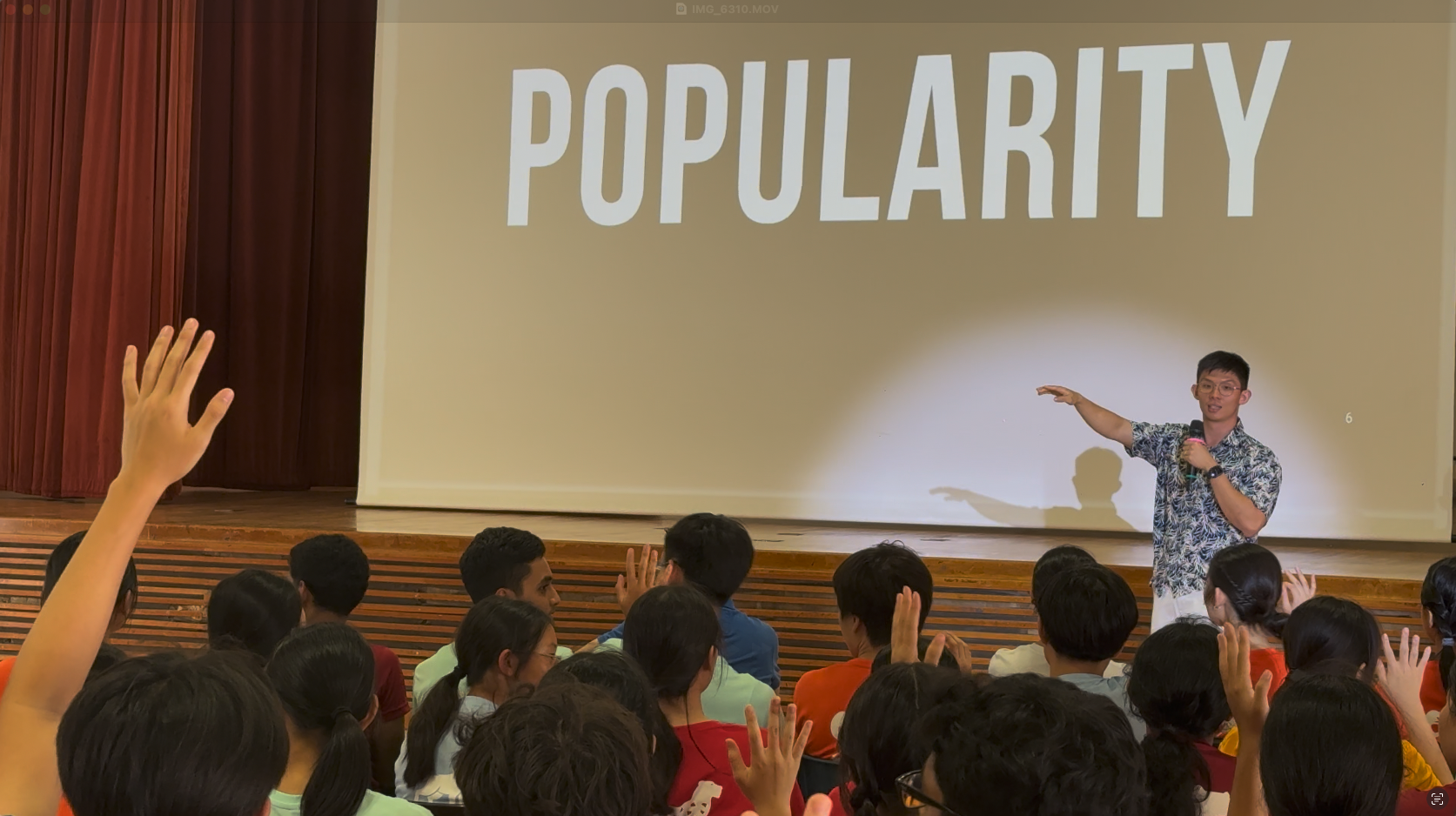 A speaker holds a microphone on stage before students raising hands, with a projection showing the word POPULARITY.