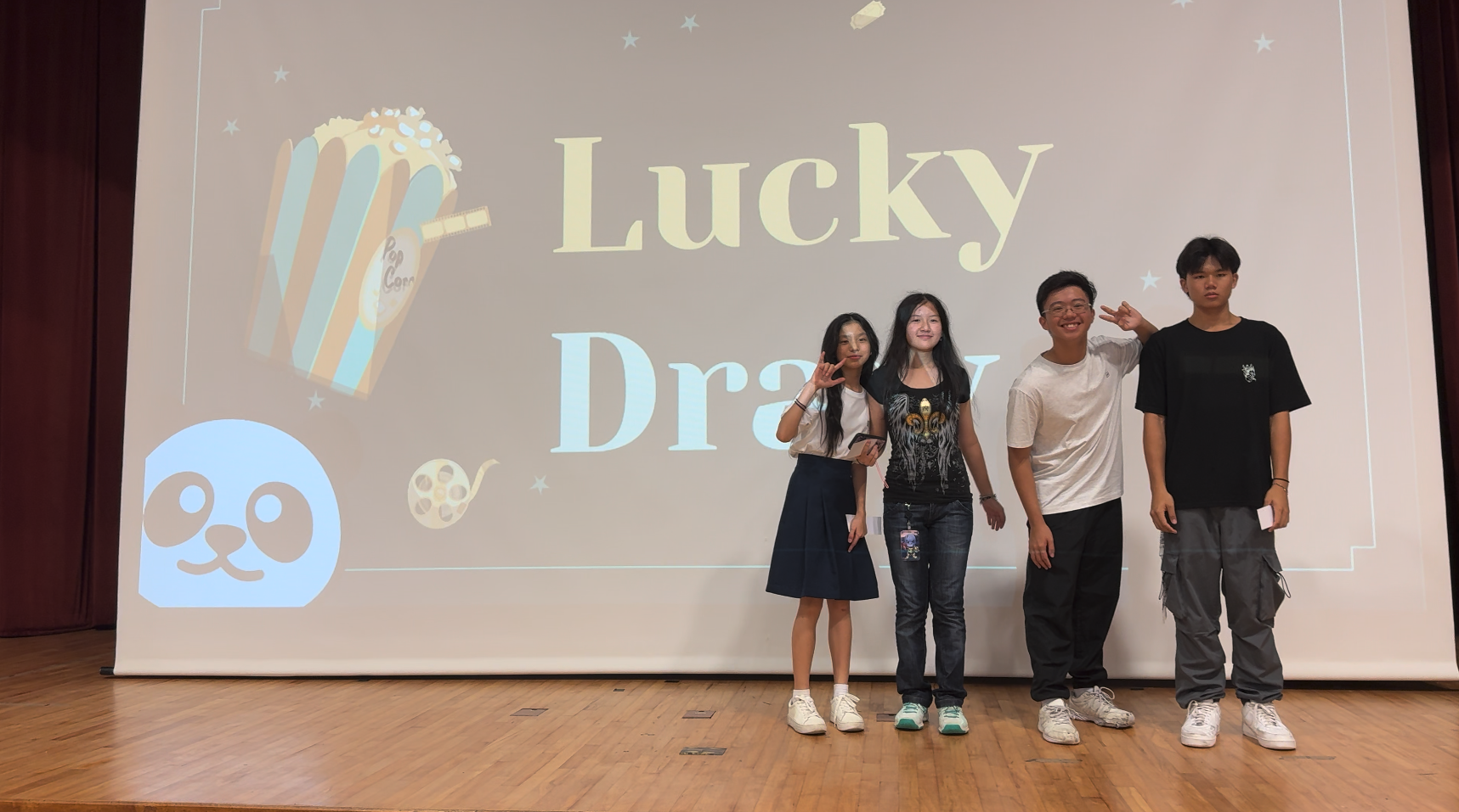 Four young people pose on a stage in front of a projected screen reading Lucky Draw with popcorn and panda graphics.